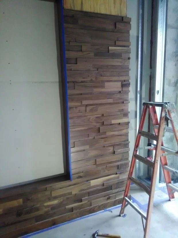 Wall with stacked wood planks being installed; orange ladder and taped unfinished wall.