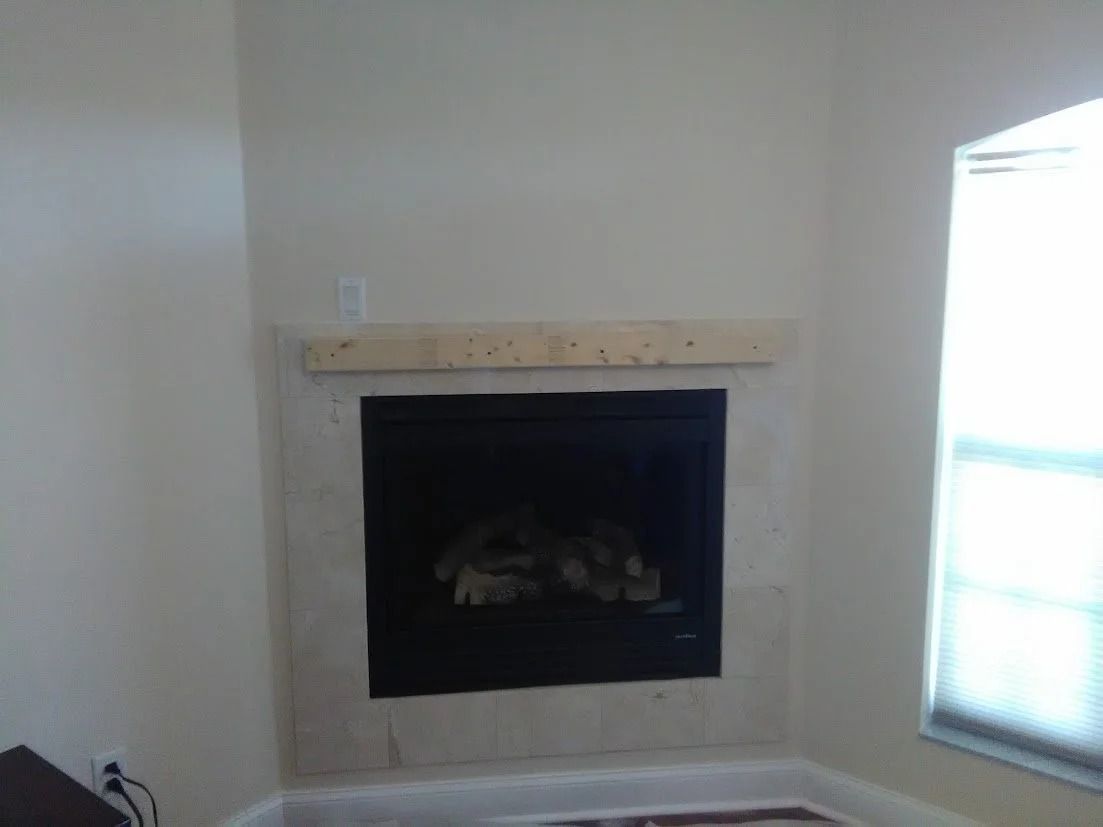 Fireplace with stone surround and wooden mantel in a room with light-colored walls and a window.