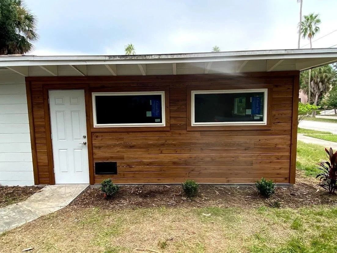 Brown wood-paneled building exterior with a white door, two windows, and a small flower bed.