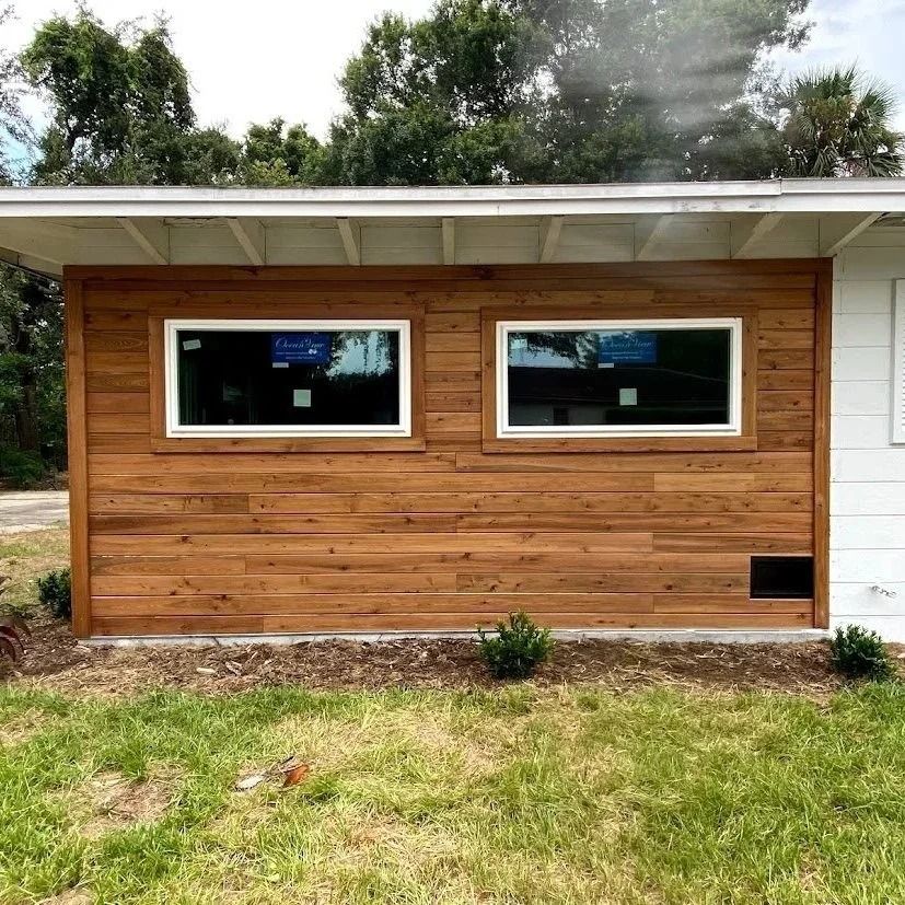 Exterior wall of a house with horizontal wood siding and two white-framed windows; green lawn below.