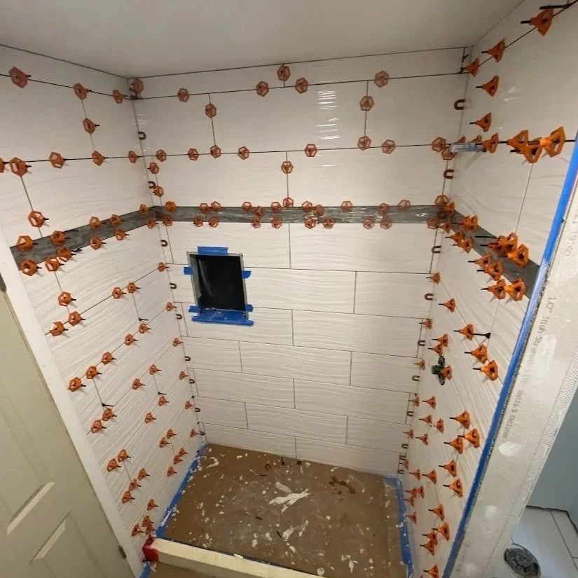 Bathroom tiled walls in progress, orange spacers, blue tape, and a small opening for a fixture.