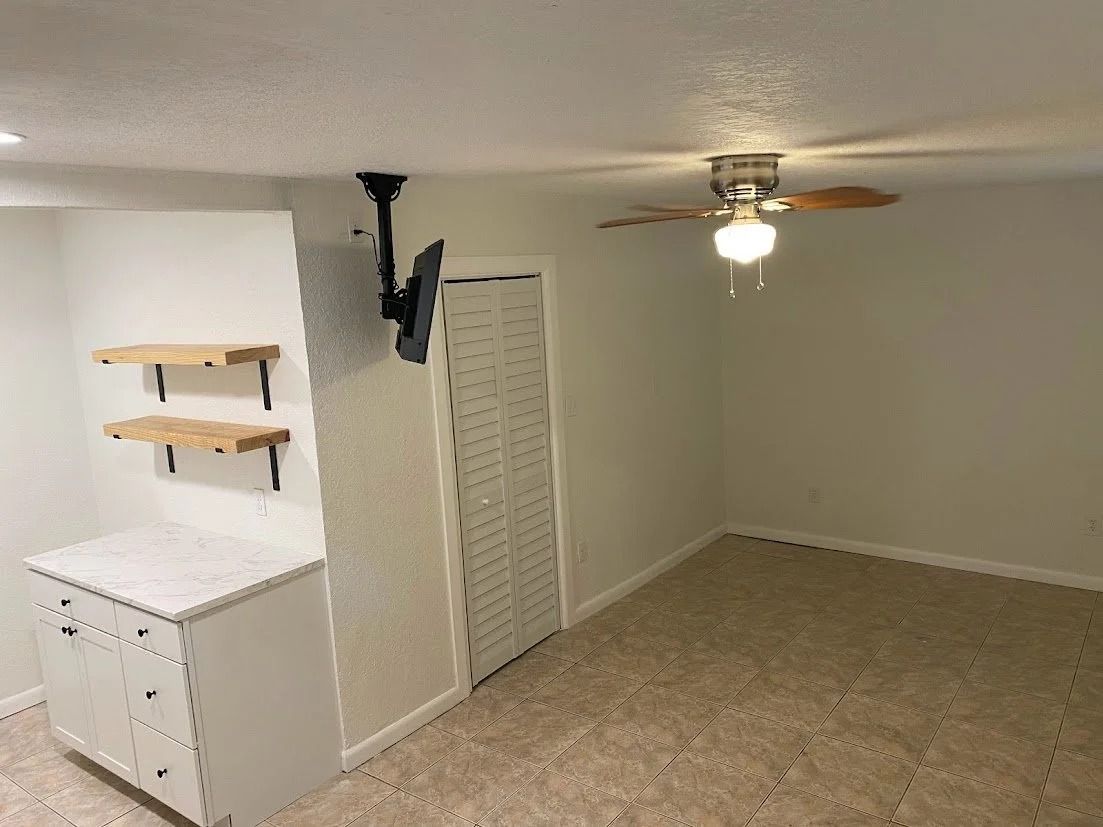 Empty bedroom with beige carpet, white walls, built-in cabinet, ceiling fan, and mounted TV.