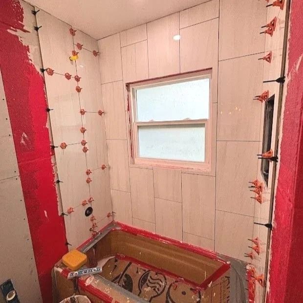 Bathroom under renovation with tile installation; red waterproofing, tiles installed around a window and tub.