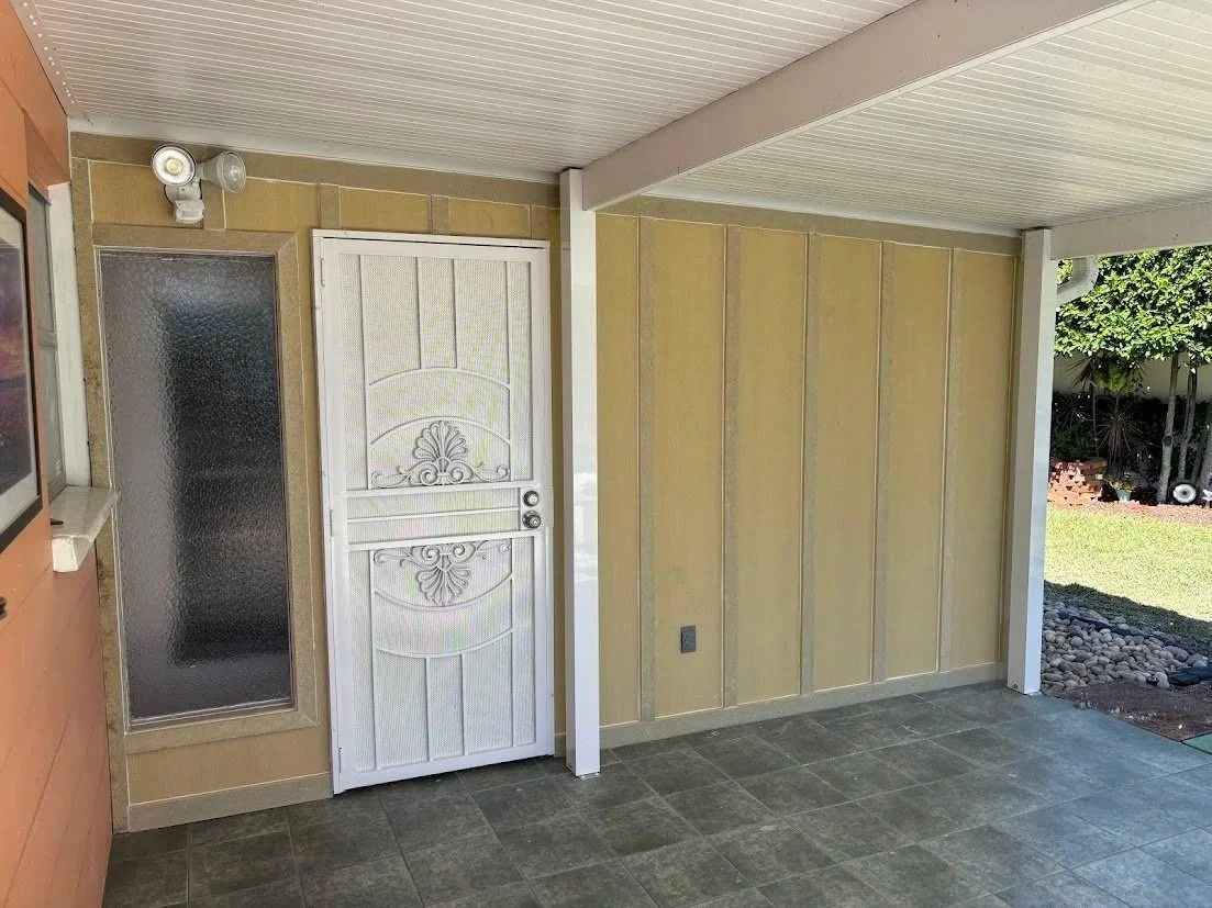 Covered patio with a screen door and security lights; light yellow walls, concrete floor, green lawn visible.
