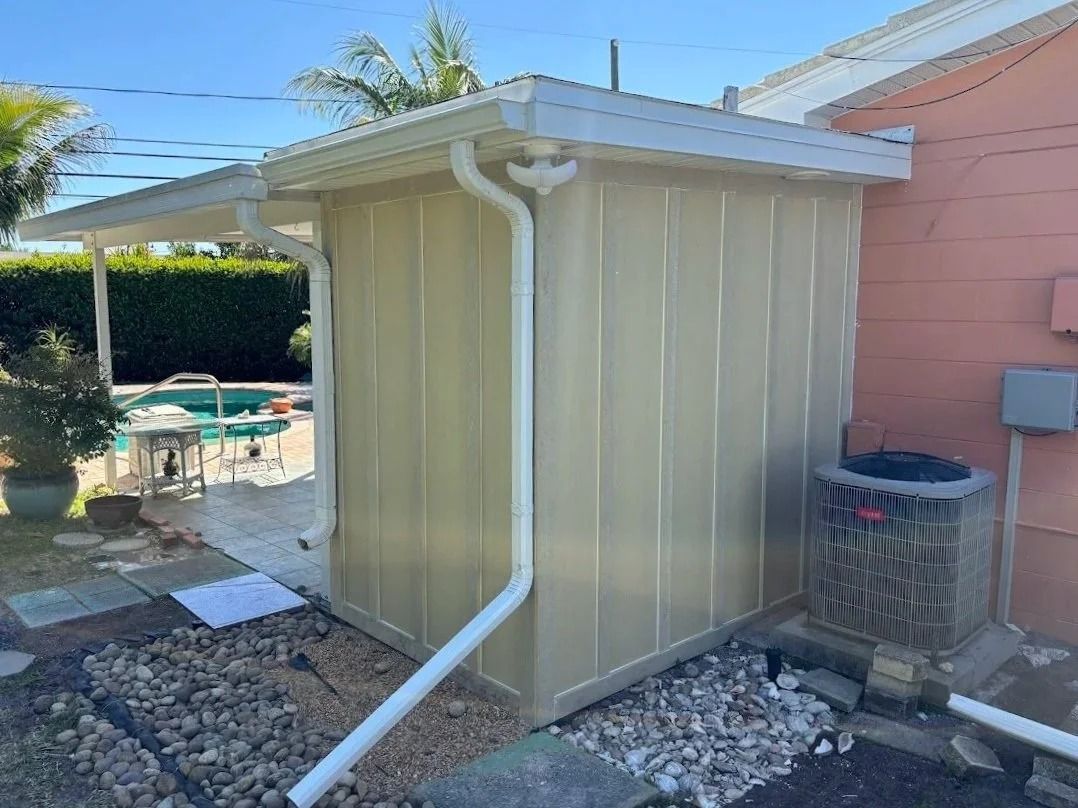 A small, beige shed with white gutters and roof is next to a pink building.  An air conditioning unit sits below the shed.