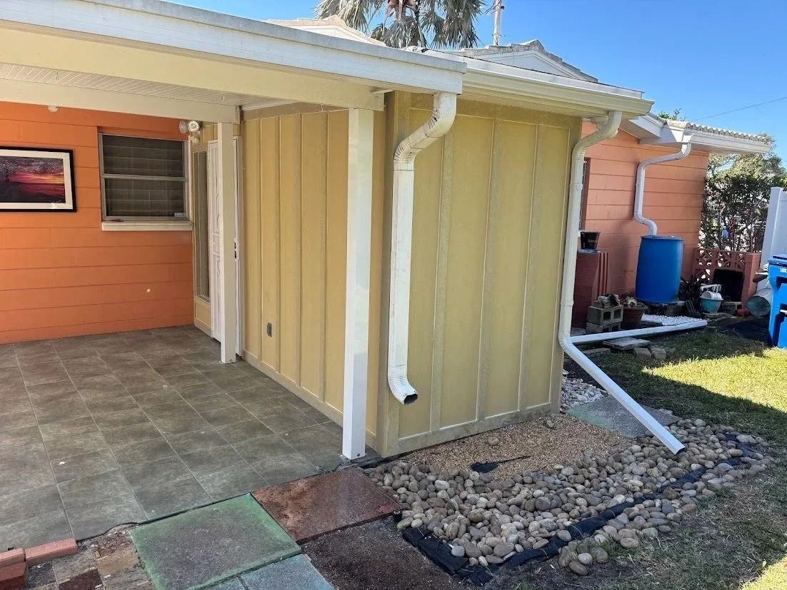 A yellow-sided shed with white gutters and roof against an orange house; a patio and gravel are in front.