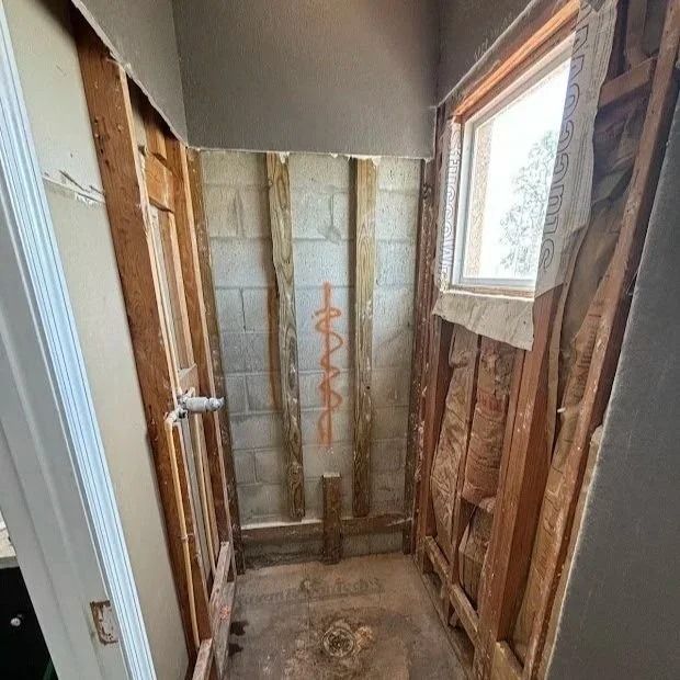Bathroom under construction: exposed framing, concrete block wall, window, and plumbing visible.