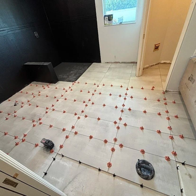 A bathroom floor with light-colored tiles, red leveling clips. Black wall and open doorway.