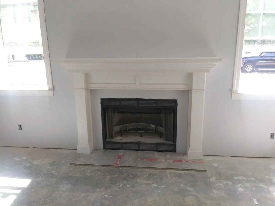 Fireplace with white mantle, flanked by windows, in a room with unfinished flooring.