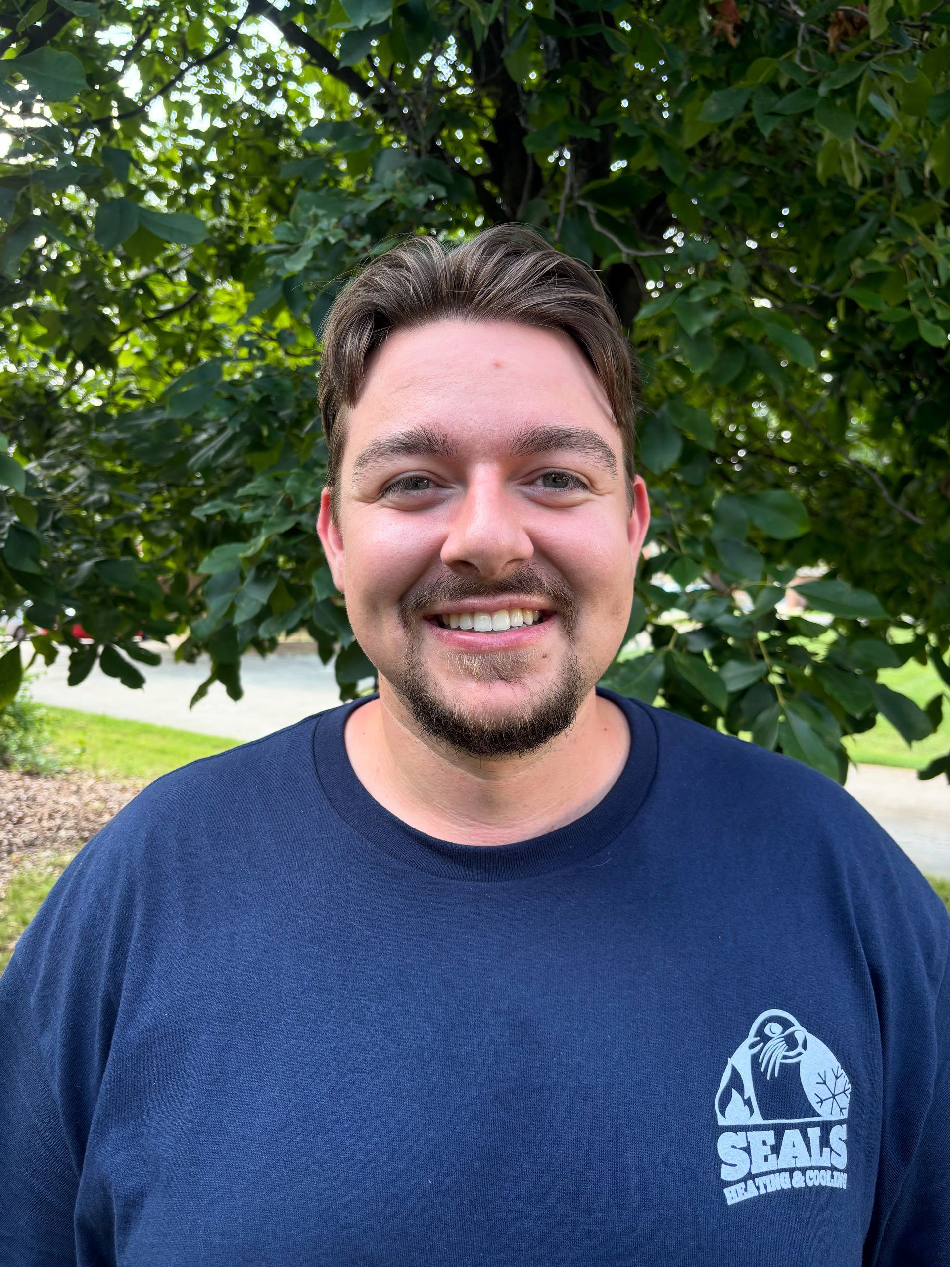 Man smiling in front of foliage, wearing a navy blue sweatshirt with a 