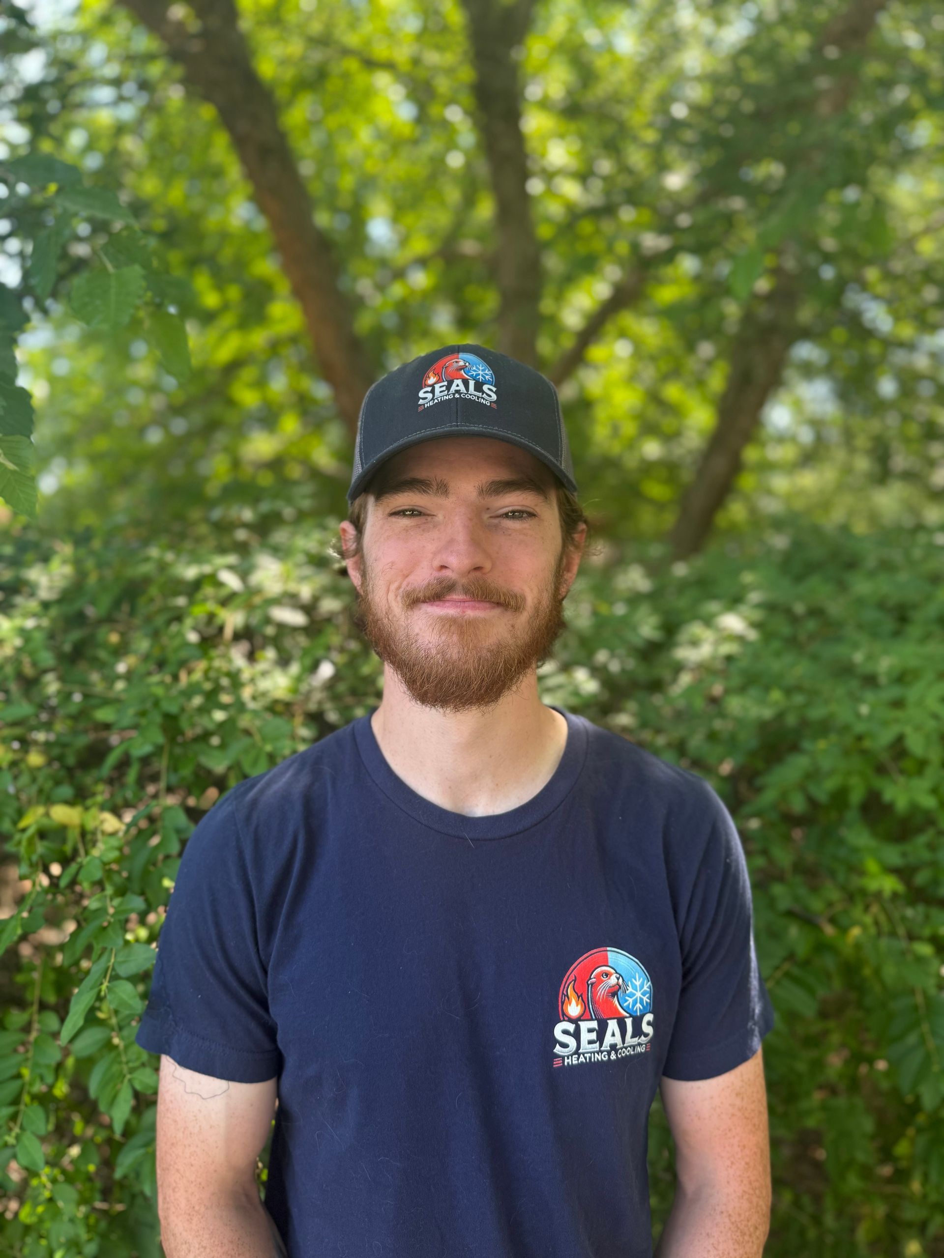 Man in blue shirt and cap with logo, smiling in front of greenery.