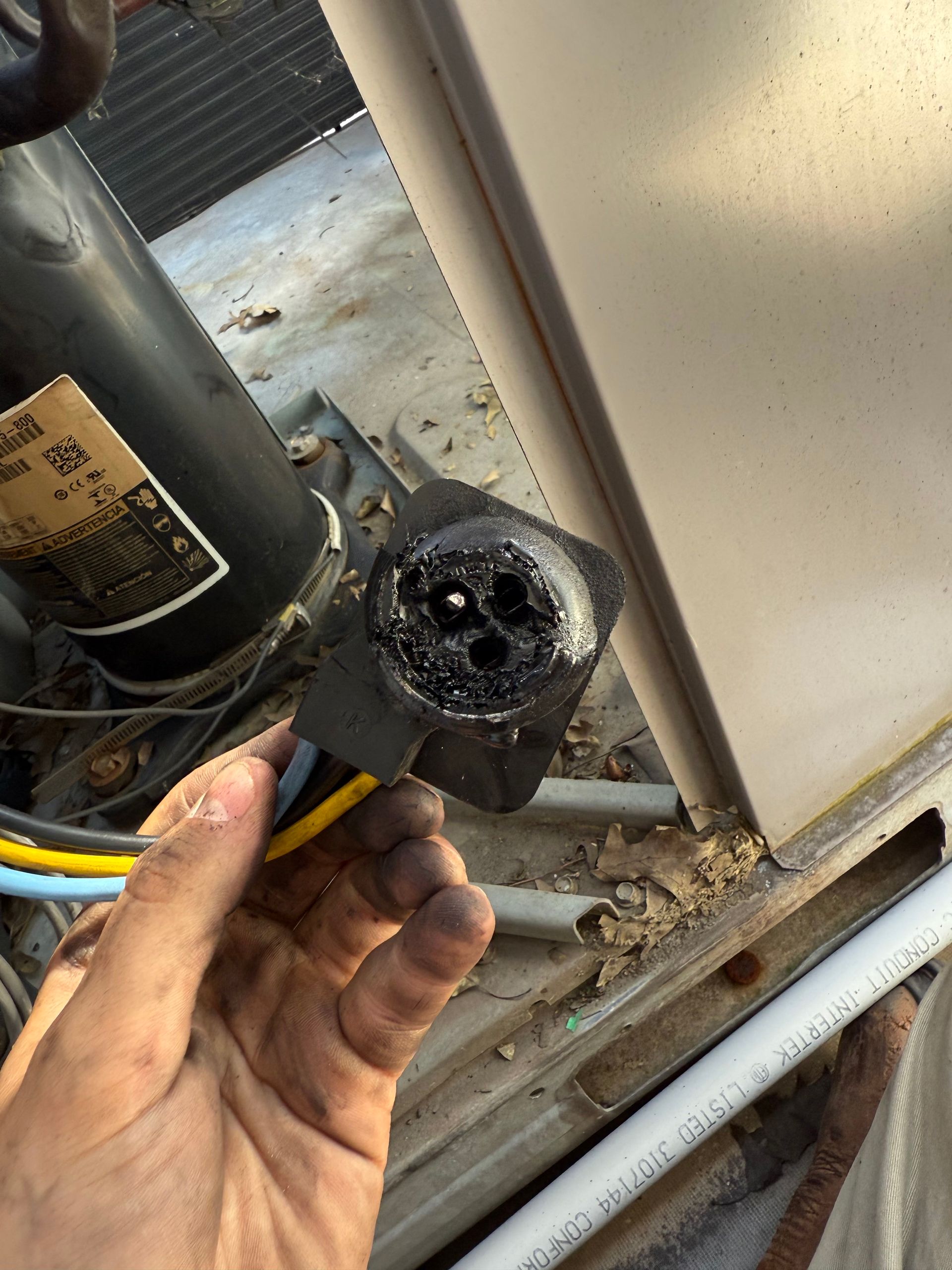 Hand holding a damaged black electrical connector with burnt wiring, near an AC unit.