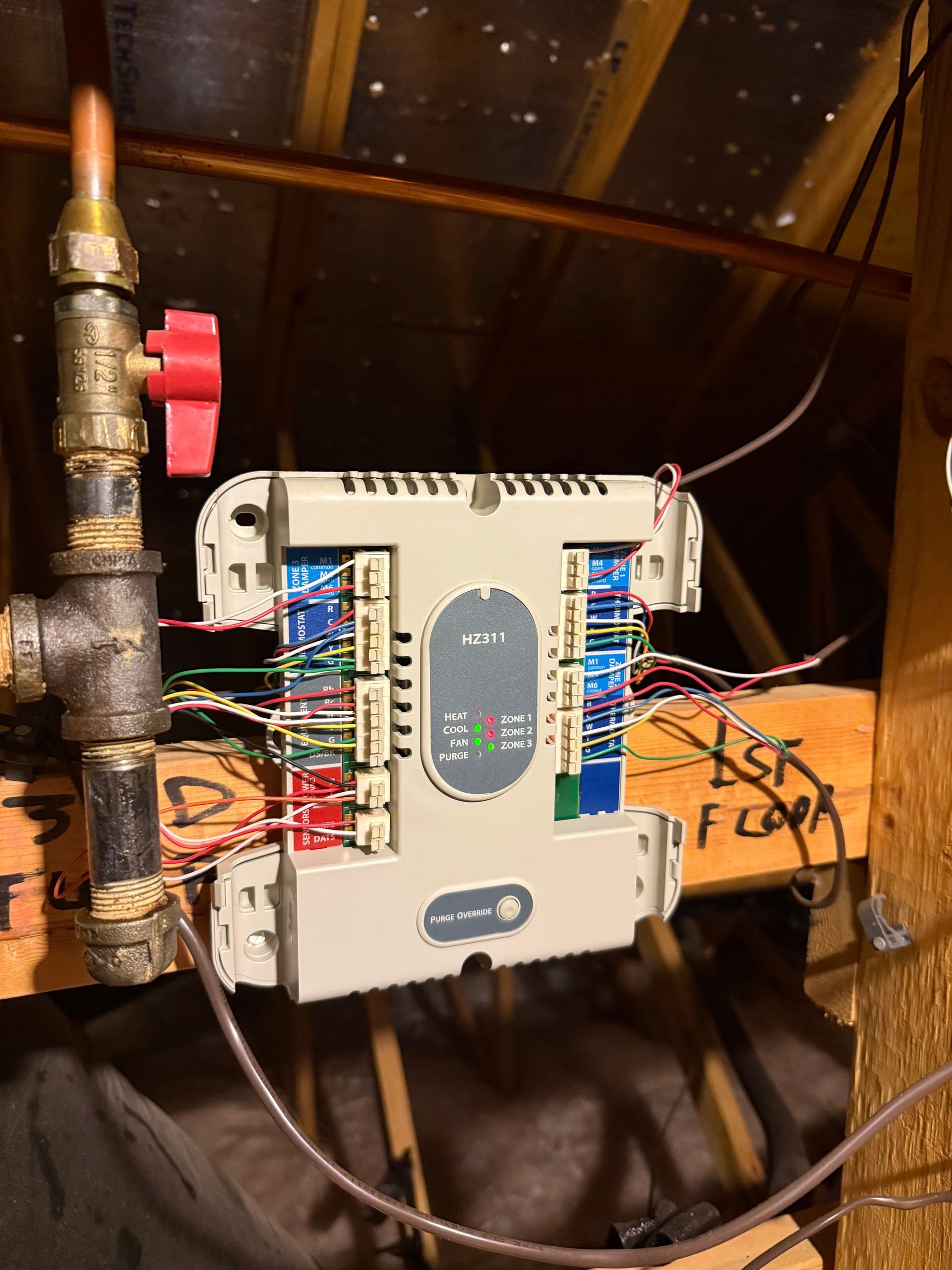 A control panel with wires, mounted on wooden rafters in an attic, next to a gas pipe with a red valve.