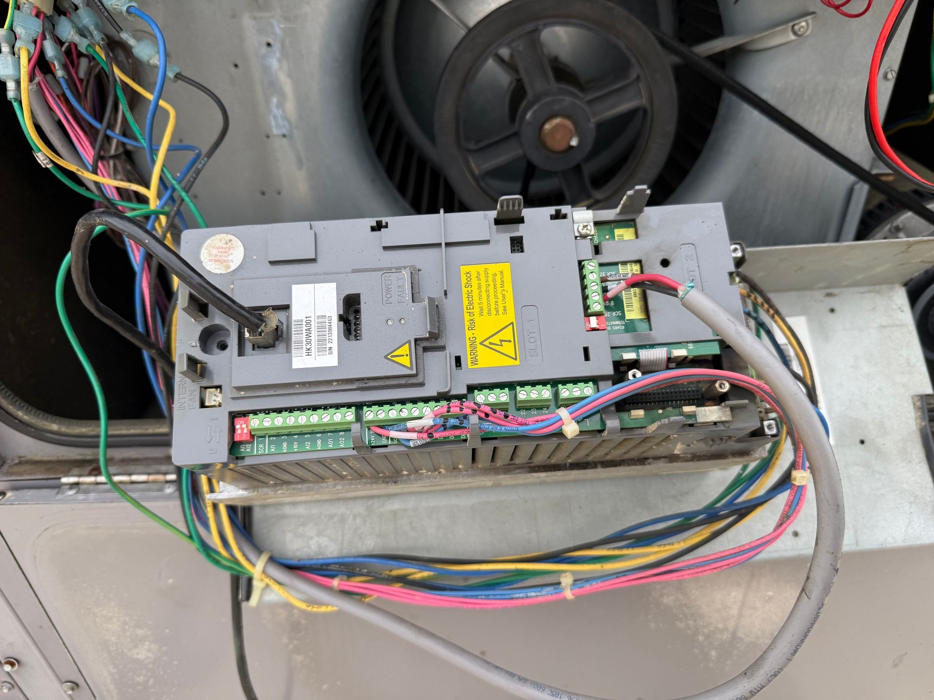 Control panel with wires, fan, and pulley. Grey panel with green terminals and yellow hazard labels.