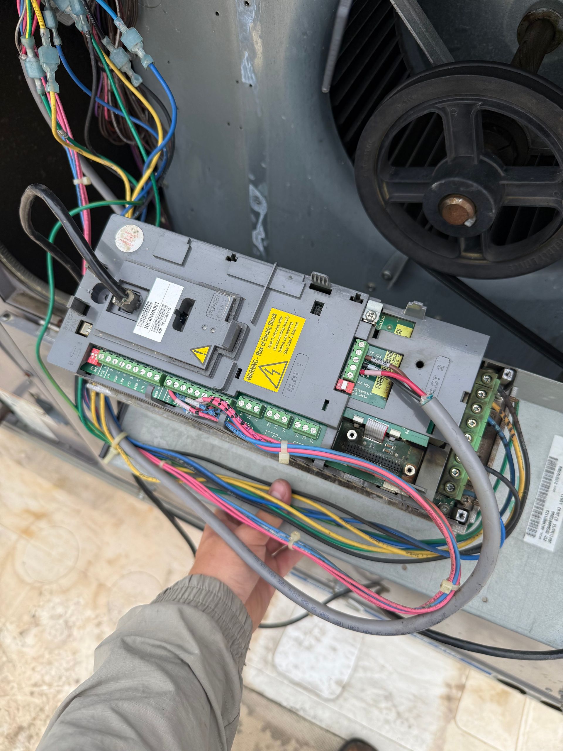 Hand holding an electrical component with colored wires connected to a large HVAC unit with a belt and pulley.