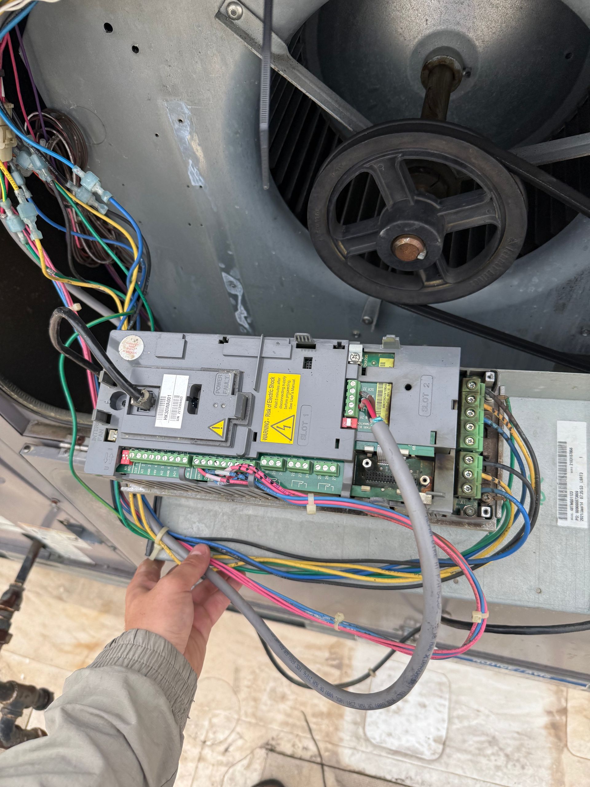 Hand holding wires connected to an HVAC control panel, near a fan and pulley system.