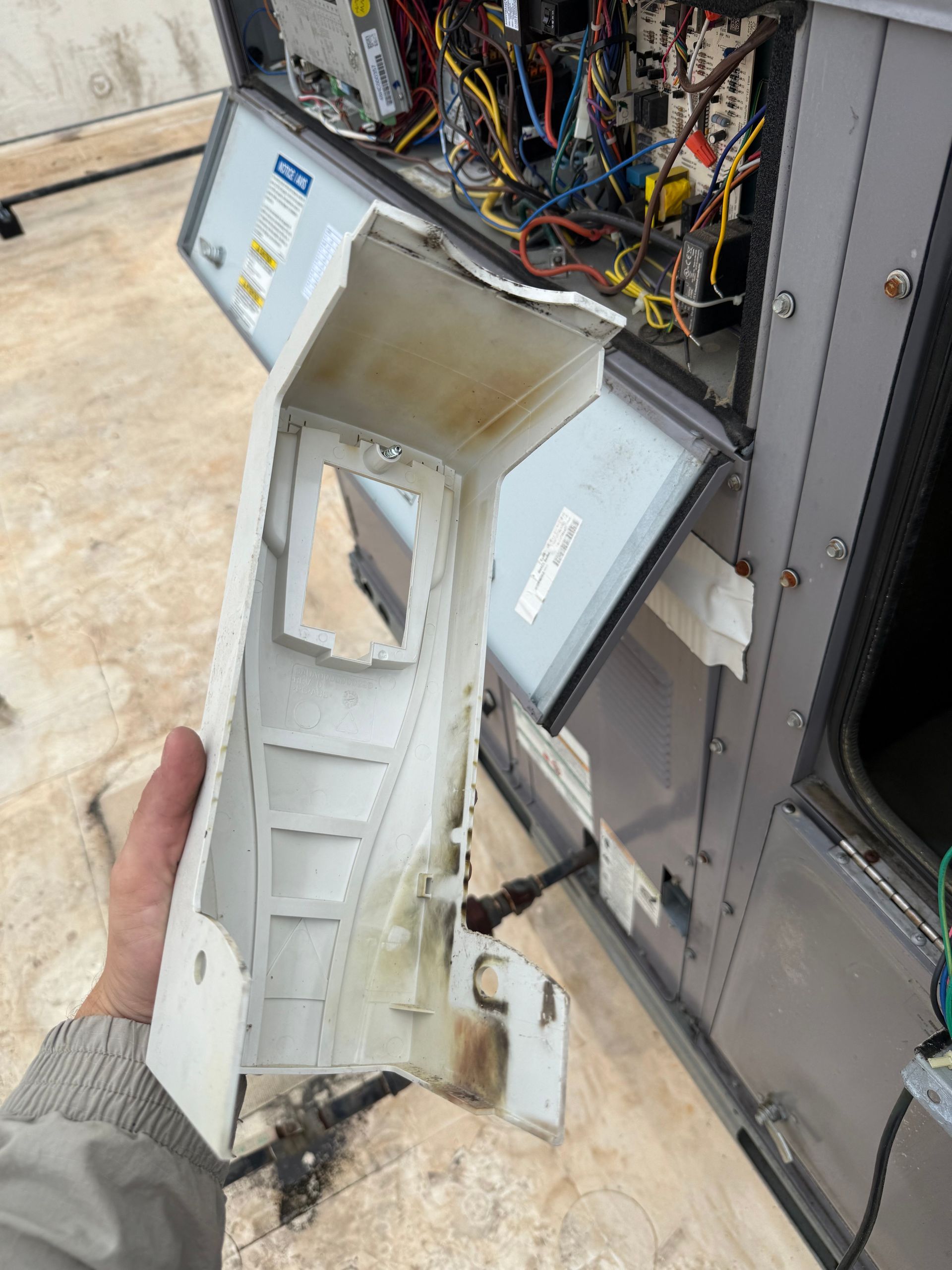 A hand holding a broken white plastic HVAC panel next to an opened unit on a rooftop.