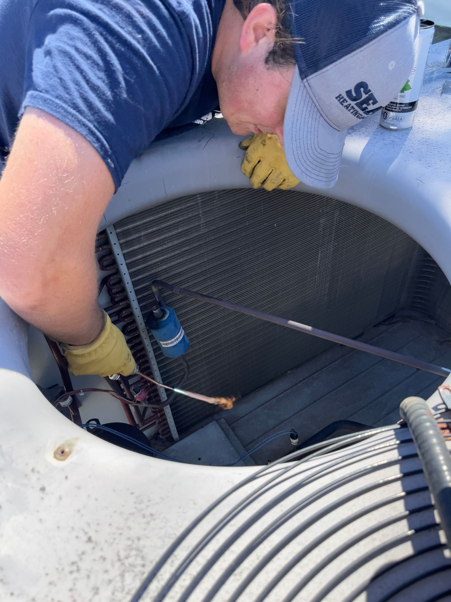 A person cleans an AC unit with a spray bottle and brush. Wearing gloves and a hat, they lean over the coils outside.