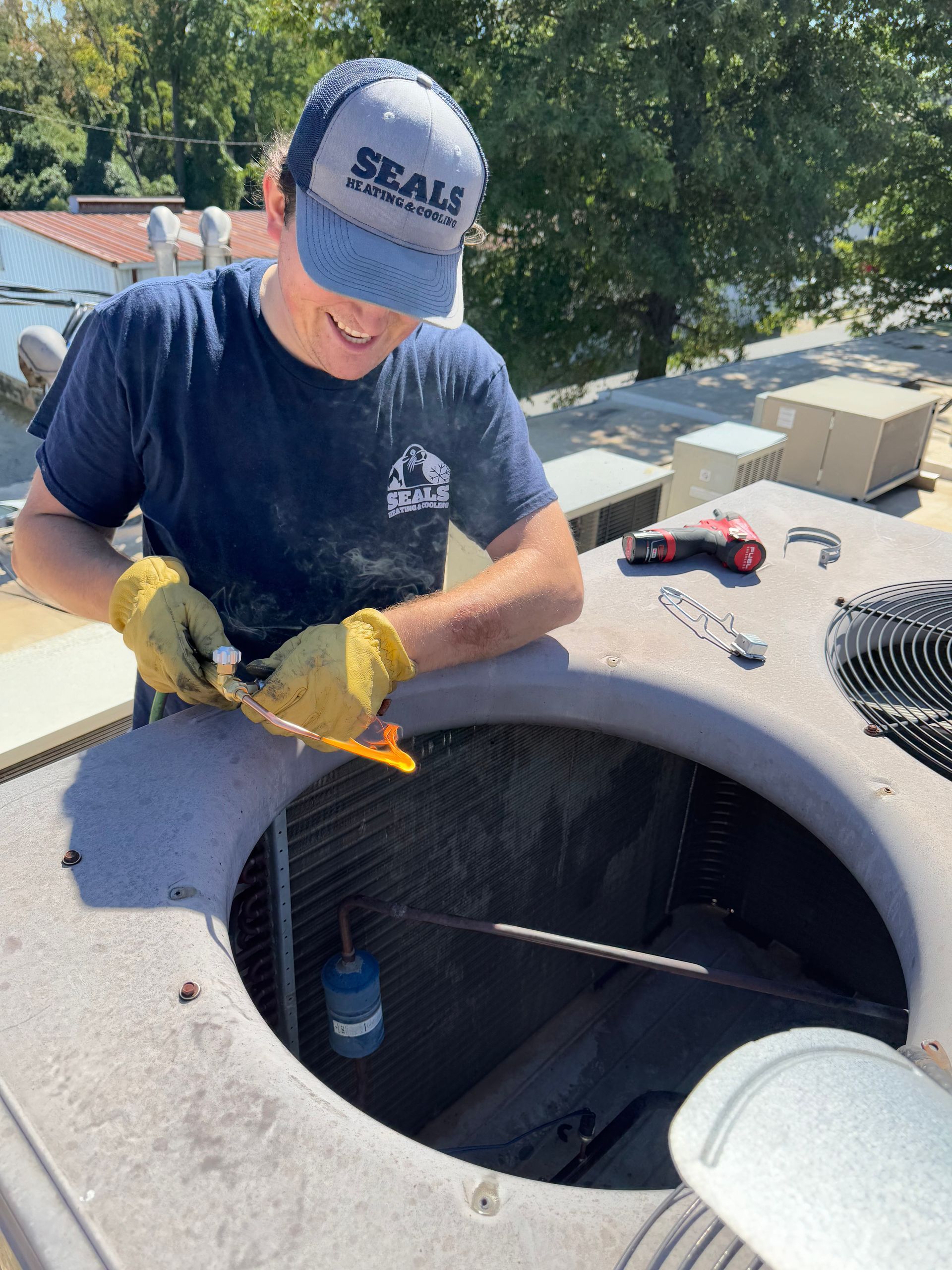 HVAC technician in a navy shirt and cap, smiling, works on an AC unit outdoors.