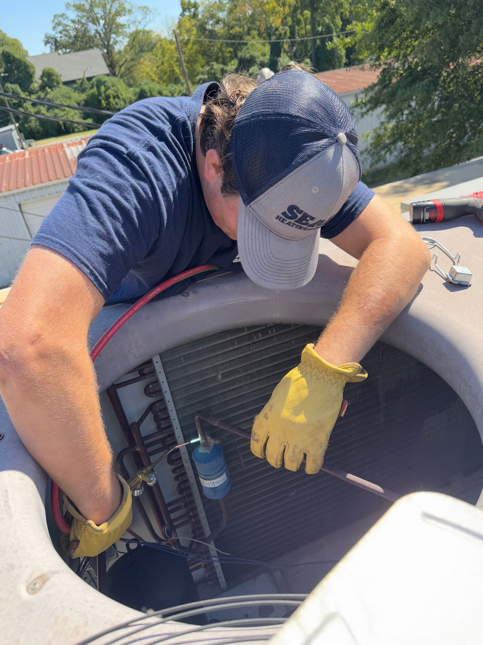 HVAC technician in a blue shirt and hat, working on rooftop air conditioning unit, wearing gloves.