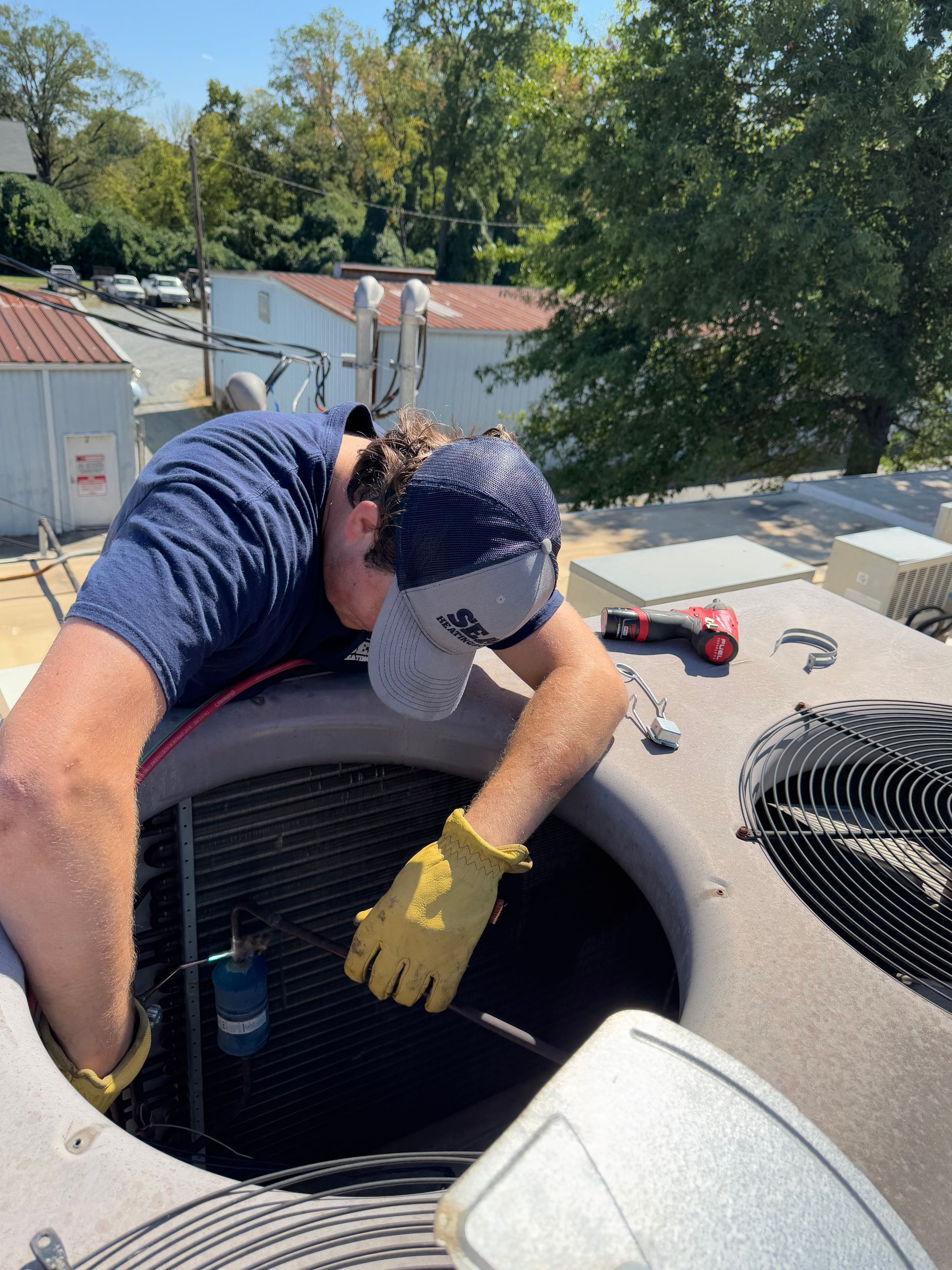 HVAC technician in a blue shirt and baseball cap working on rooftop unit, wearing yellow gloves.