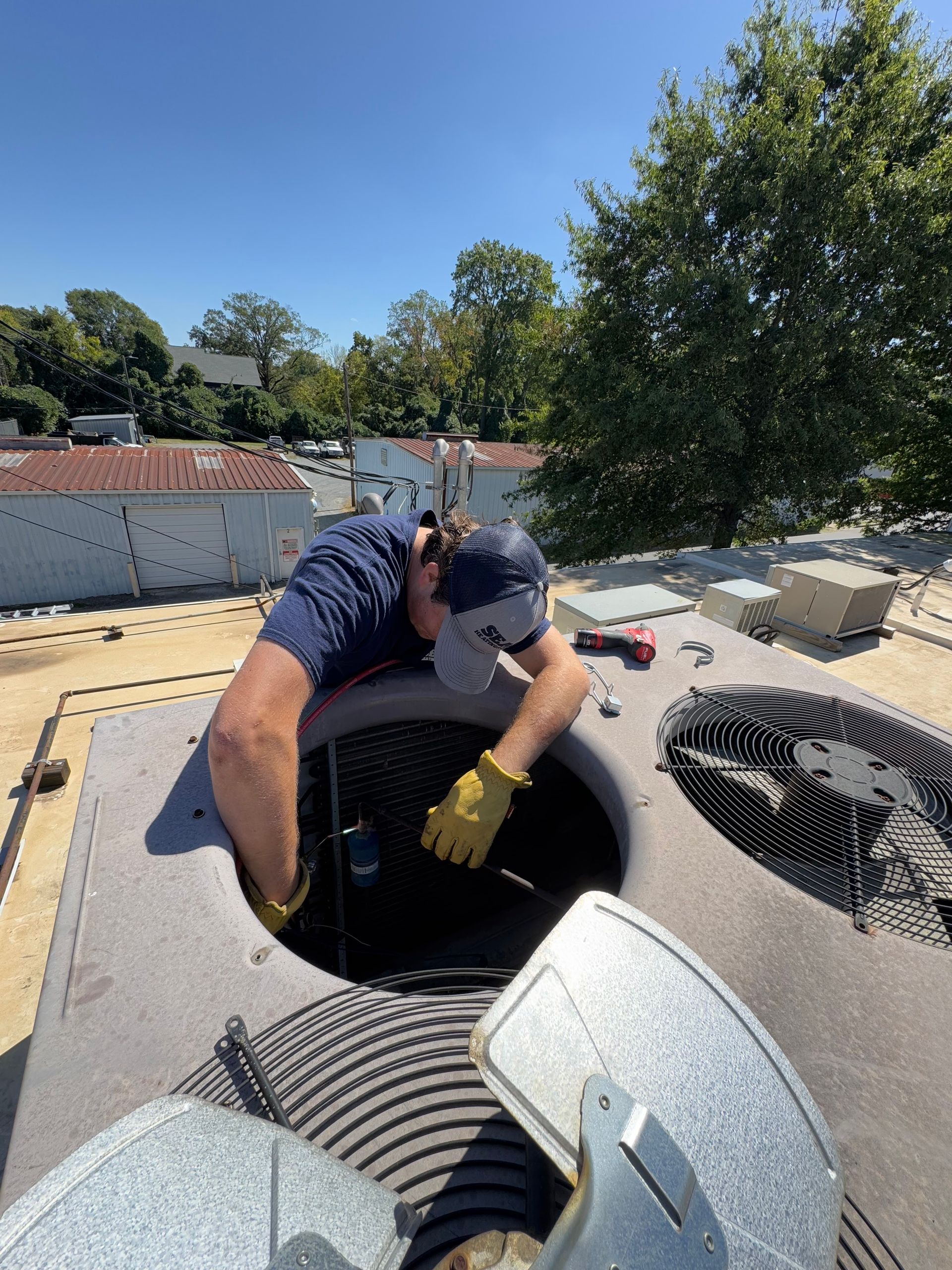 Man in blue shirt and hat working on HVAC unit on a rooftop on a sunny day.