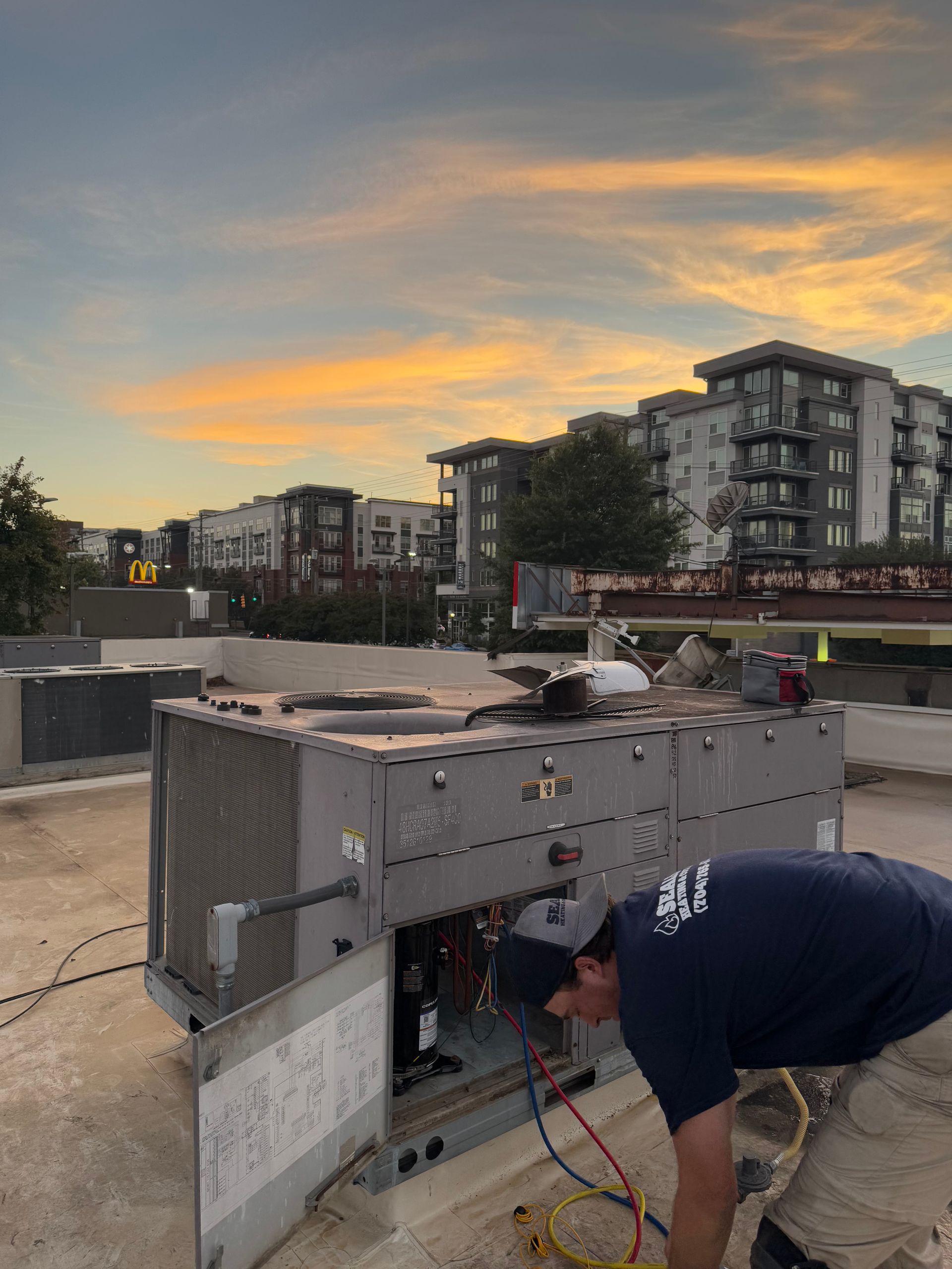 HVAC technician working on rooftop unit, sunset sky, multi-story buildings in background.
