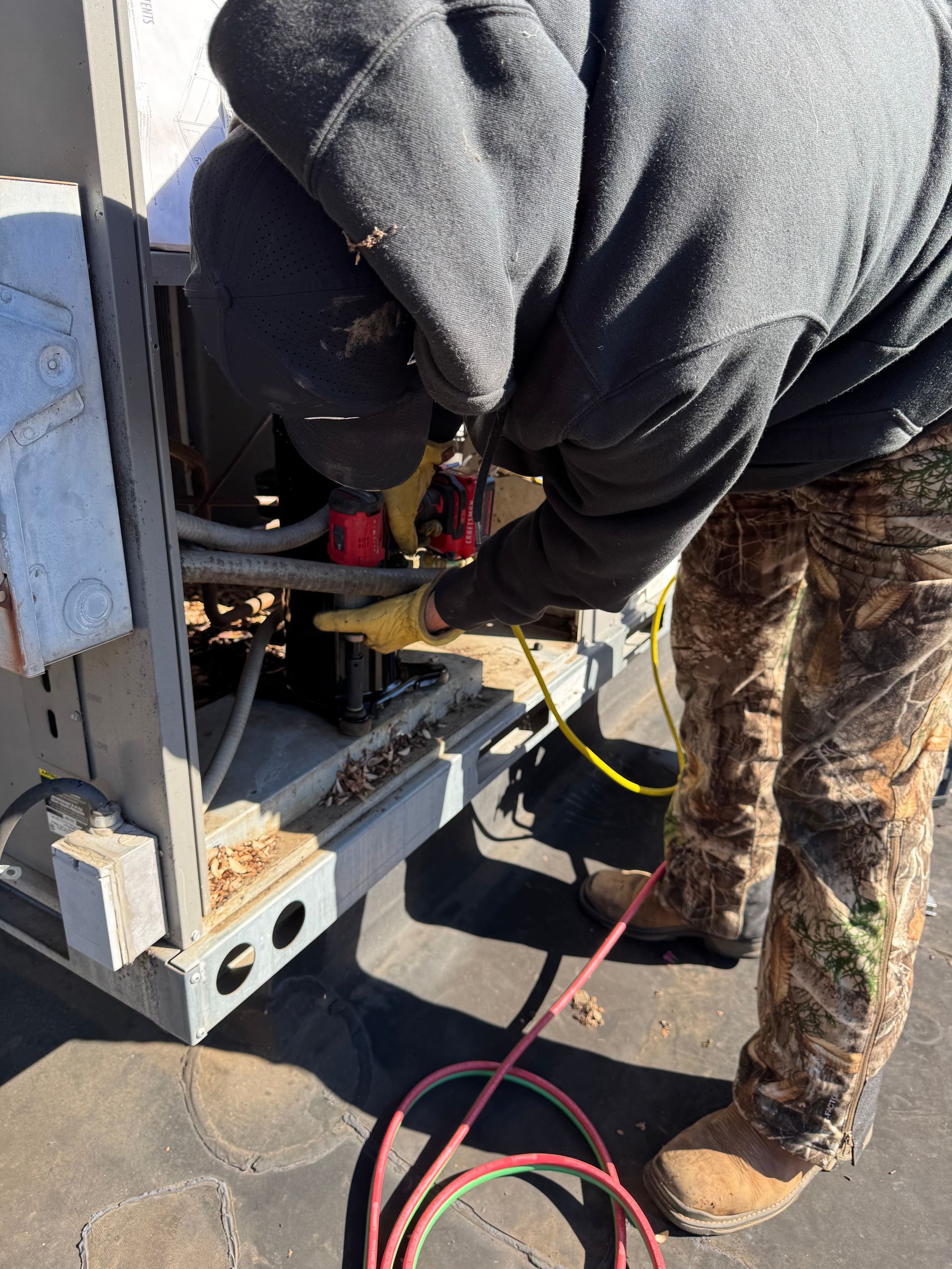 Person in hooded sweatshirt and camouflage pants working on an air conditioning unit outside.