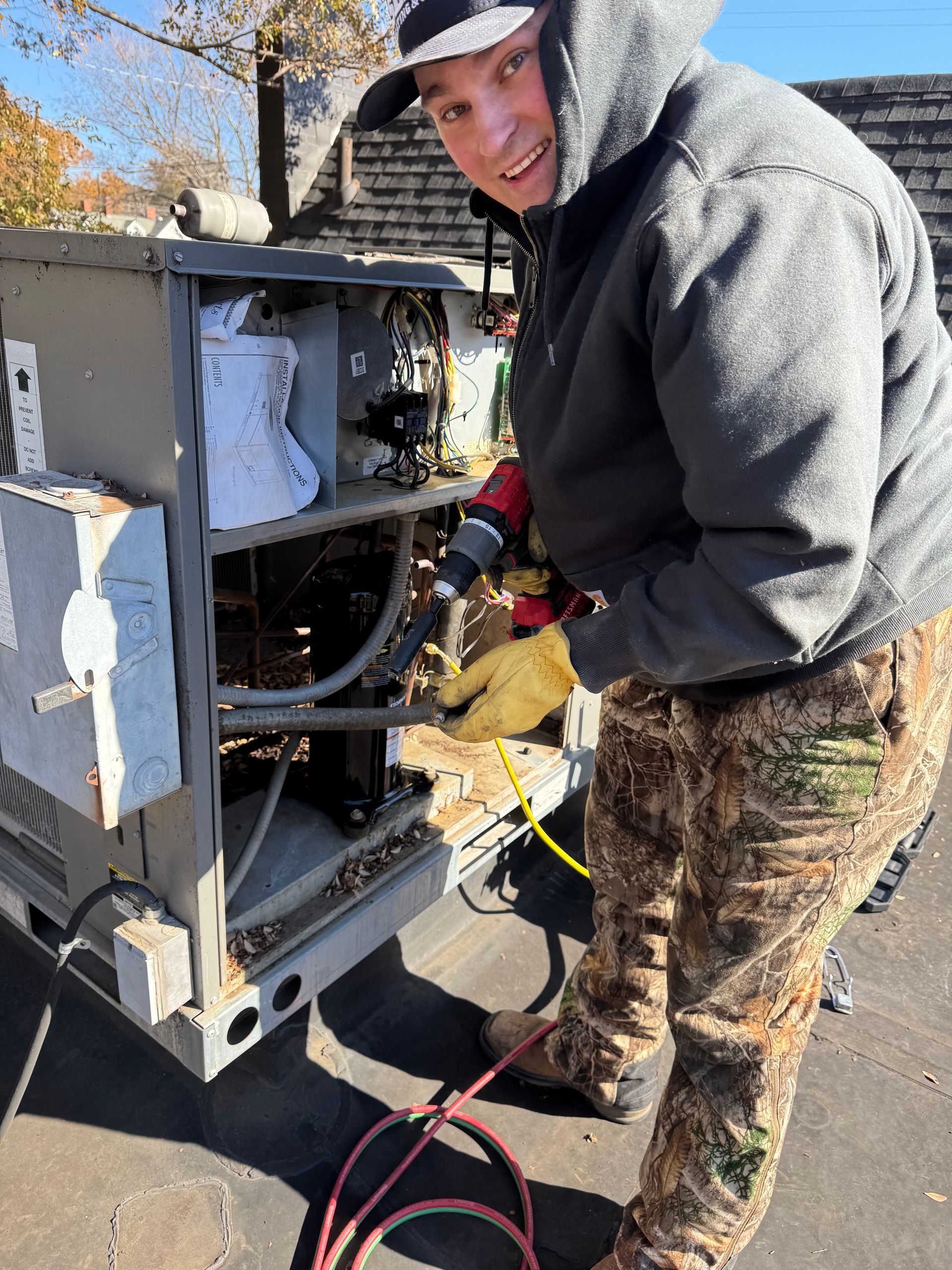 Man in camo pants, working on a machine, using a drill. He wears a hooded sweatshirt and gloves. Outdoors.