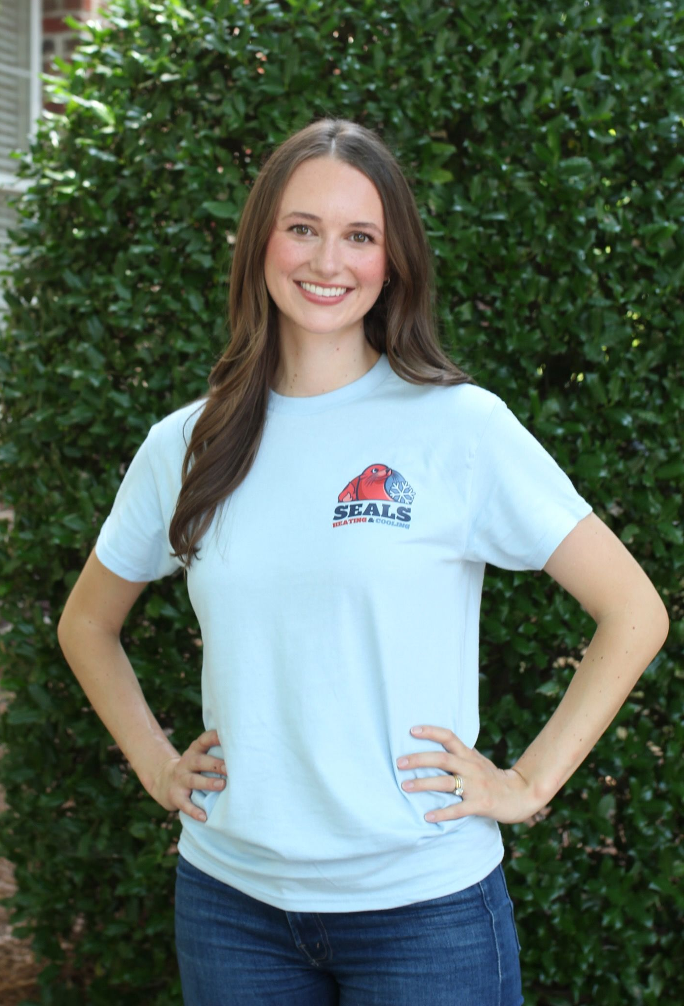 Woman in light blue t-shirt with logo, hands on hips, smiling, in front of green foliage.