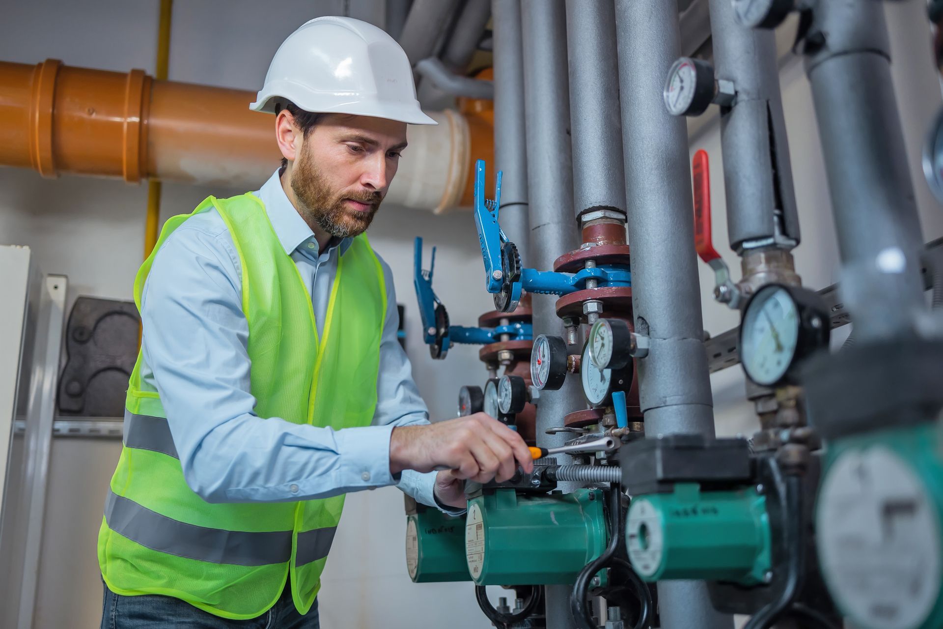 HVAC technician adjusting industrial boiler pumps and valves in a mechanical room wearing safety gear.