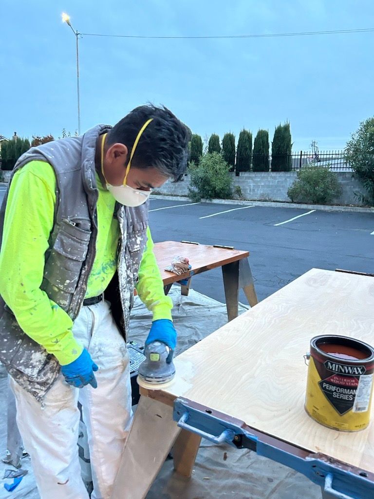 A worker wearing a face mask and safety gear sands a wooden board on a sawhorse outdoors next to a paint can.