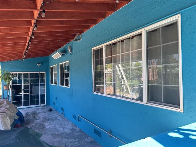 Exterior view of a bright blue wall under a wooden patio cover, featuring large windows and a glass sliding door.