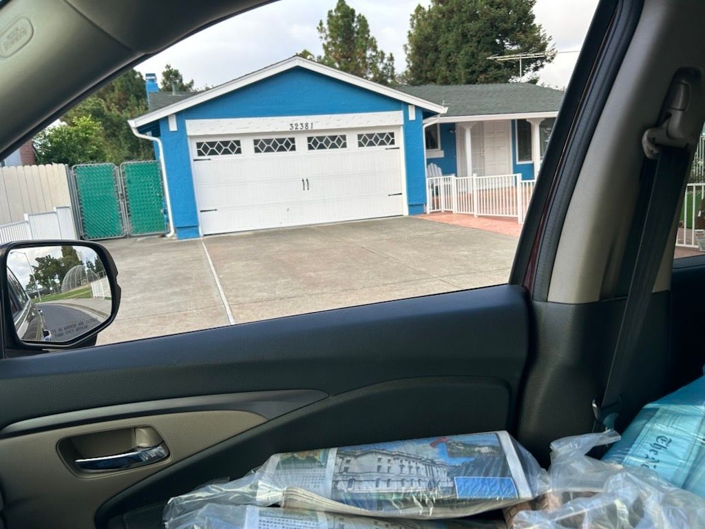 A view from inside a car looking at a bright blue house with a white garage door and fence, taken on a cloudy day.