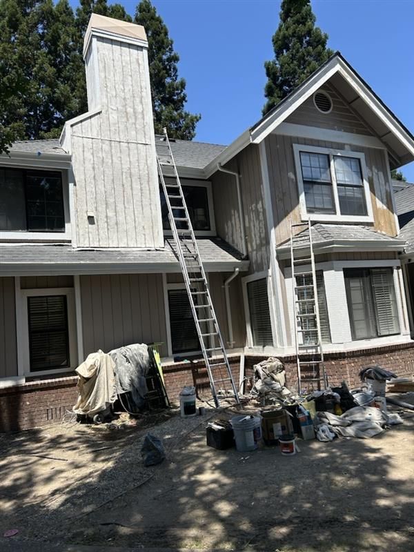 Two ladders lean against the exterior of a light brown house with a white chimney during a painting project.
