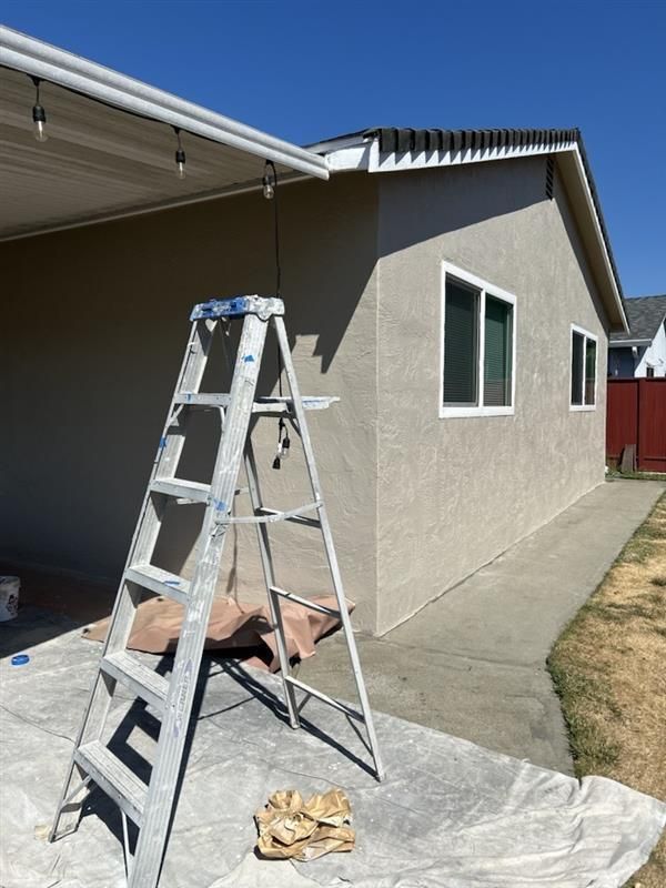 A metal step ladder sits on a drop cloth outside a beige house with a concrete walkway under a clear blue sky.