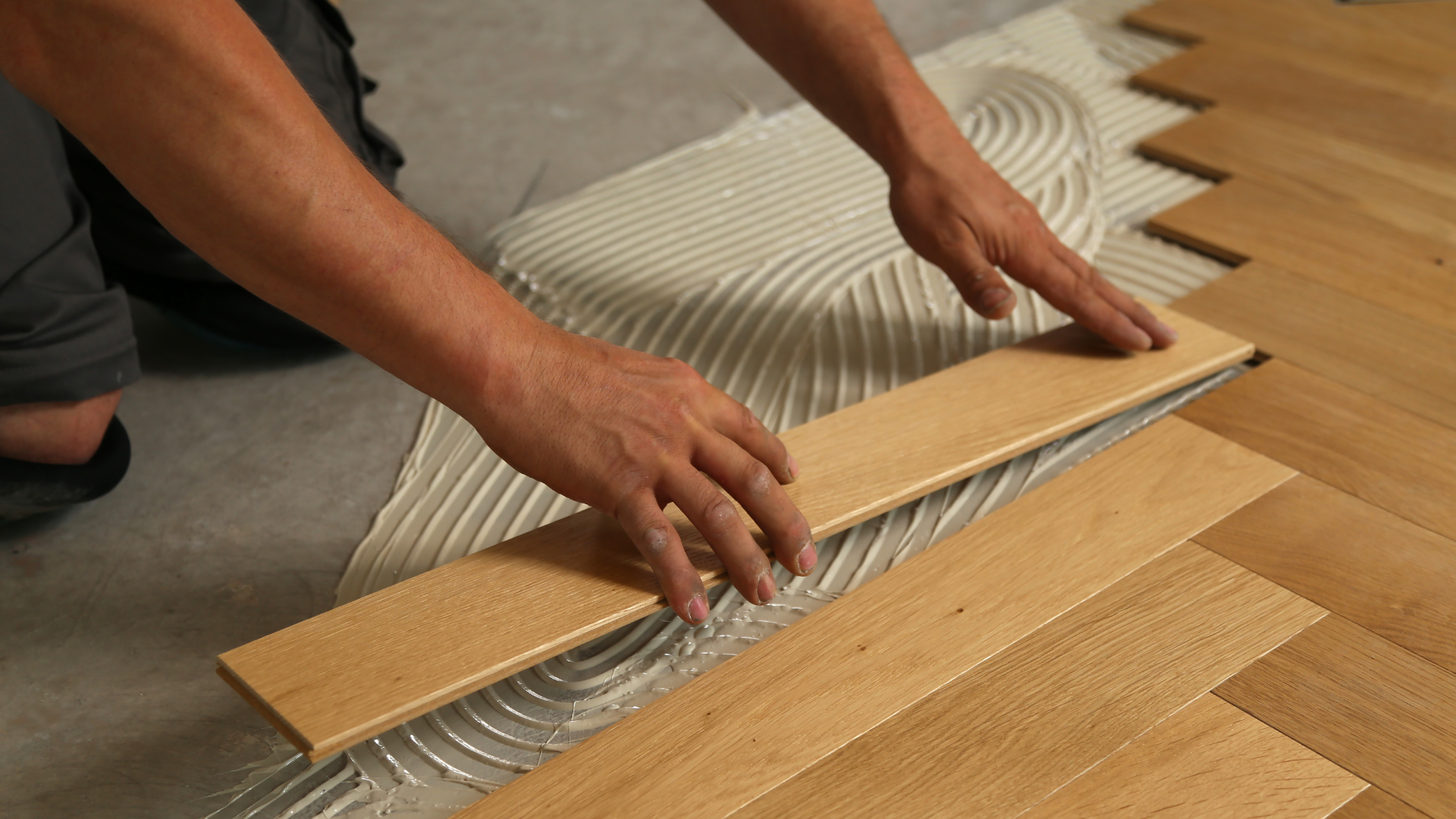 Deux mains installent des planches de bois clair en motif à chevrons sur un sol recouvert de rainures adhésives.