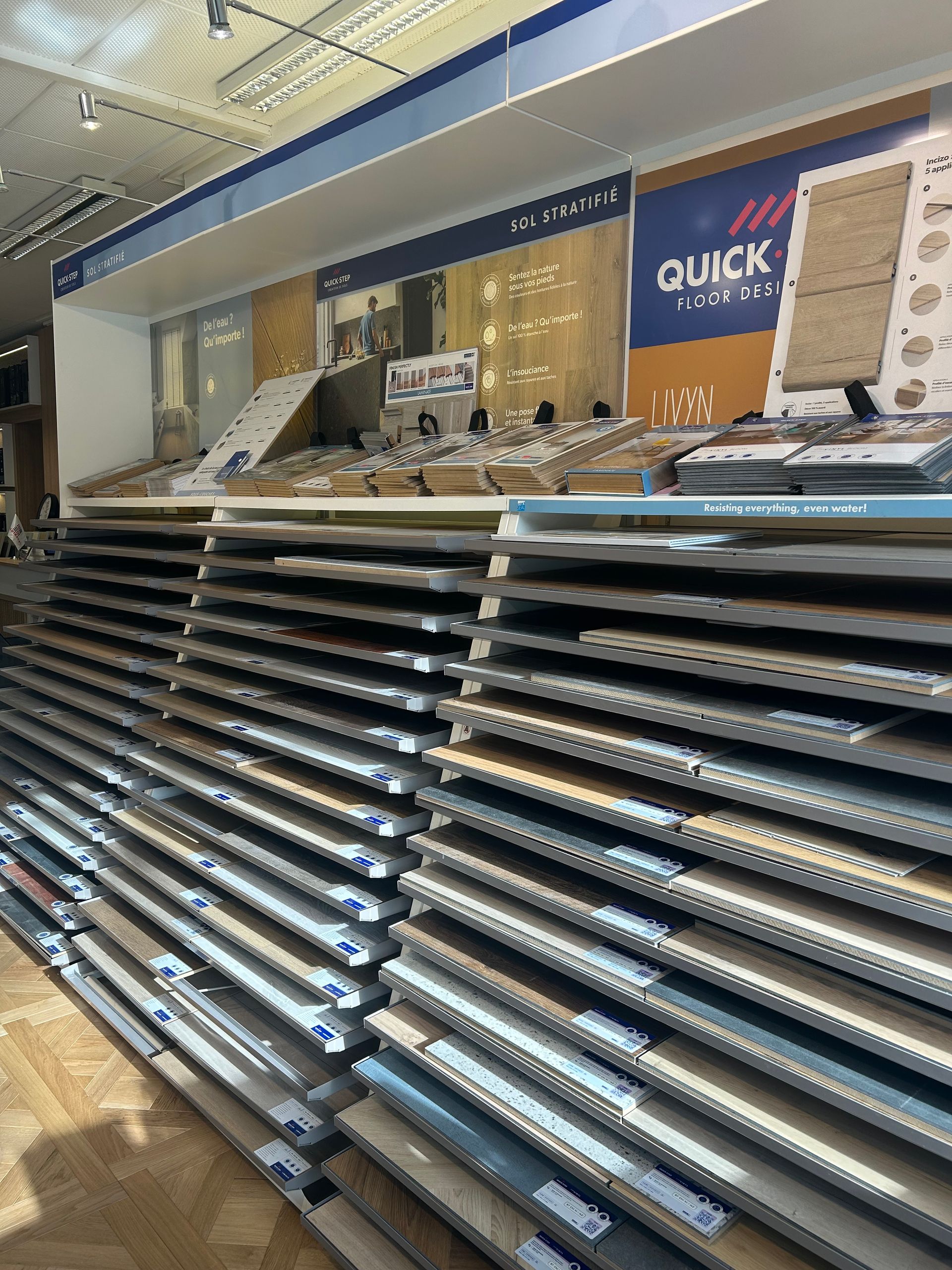 Rayonnages d'un magasin de revêtements de sol présentant des rangées d'échantillons de lames de parquet stratifié et des publicités pour ces produits.