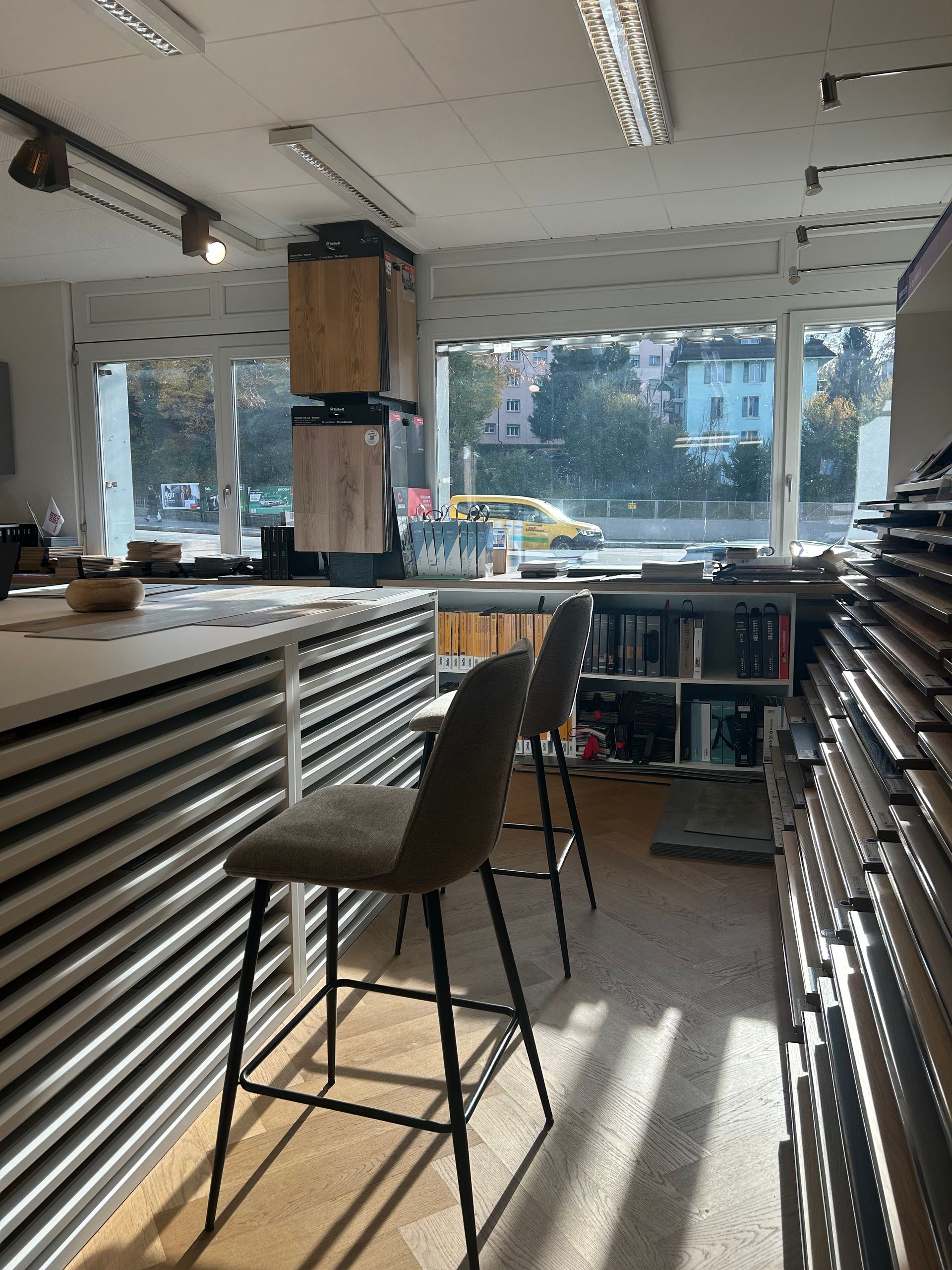 Deux tabourets de bar sont assis à un comptoir blanc dans une boutique aux grandes fenêtres donnant sur une rue et des bâtiments.