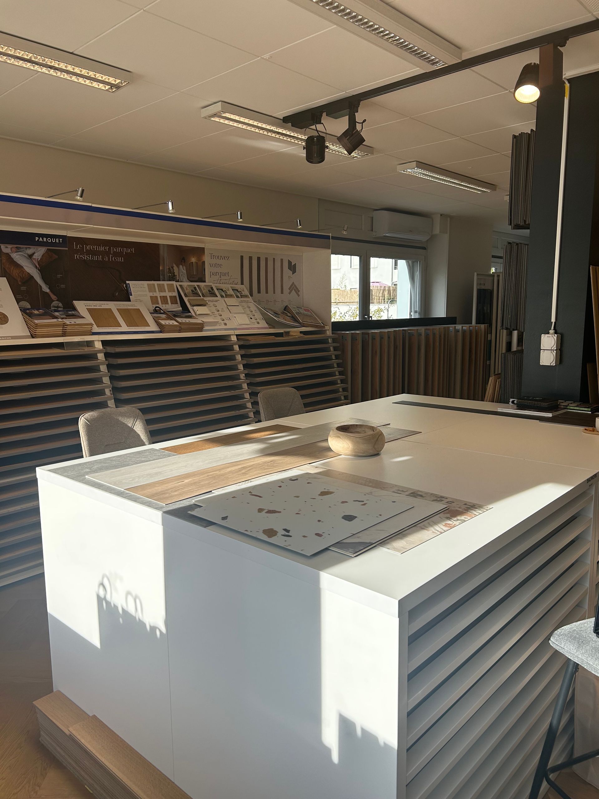 Une salle d'exposition avec une grande table centrale blanche présentant des échantillons de revêtements de sol devant des présentoirs de carrelage et de stratifié.