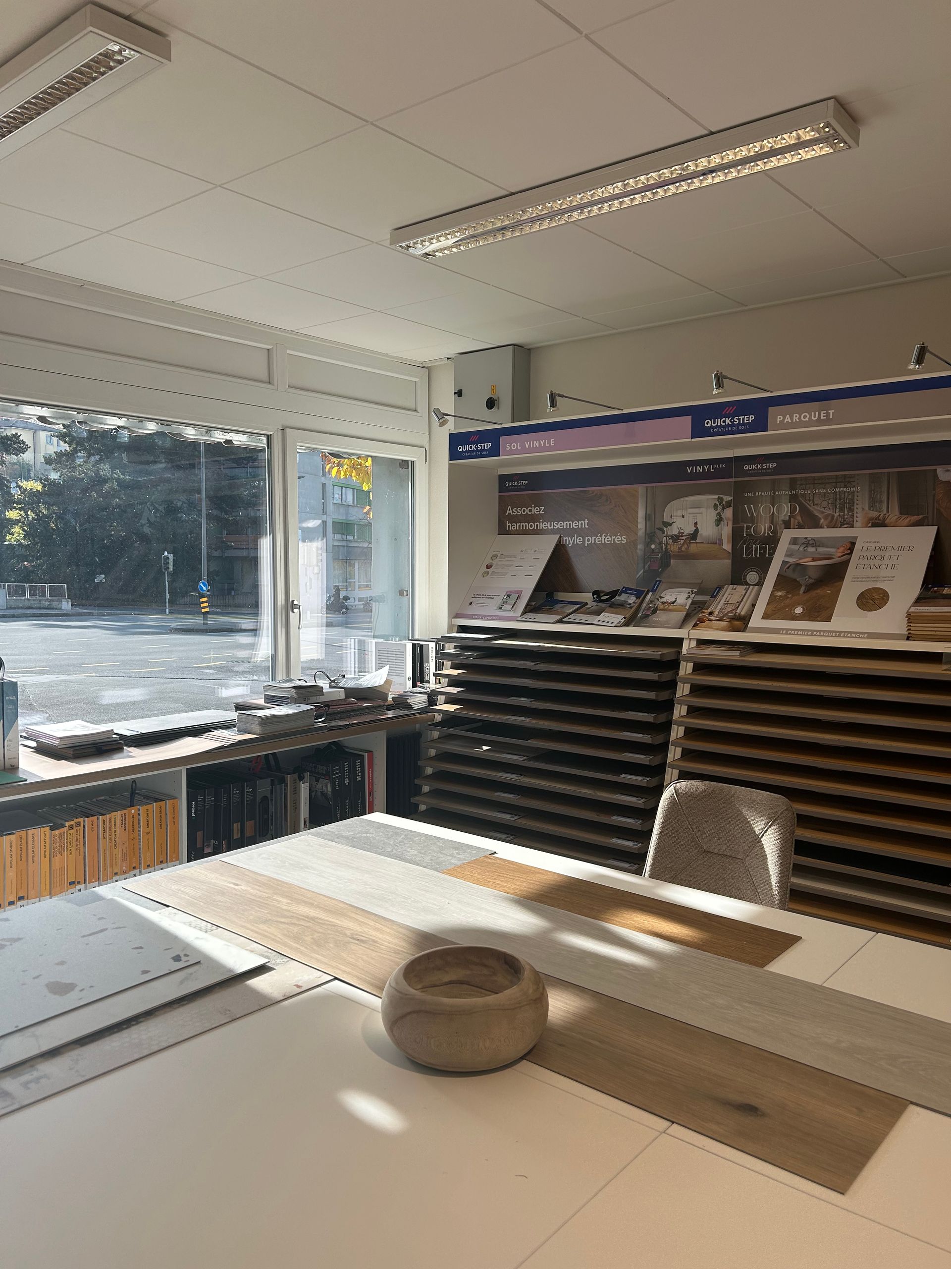 Une salle d'exposition lumineuse avec une grande table centrale présentant des échantillons de bois, des présentoirs de revêtements de sol et une grande vitrine.