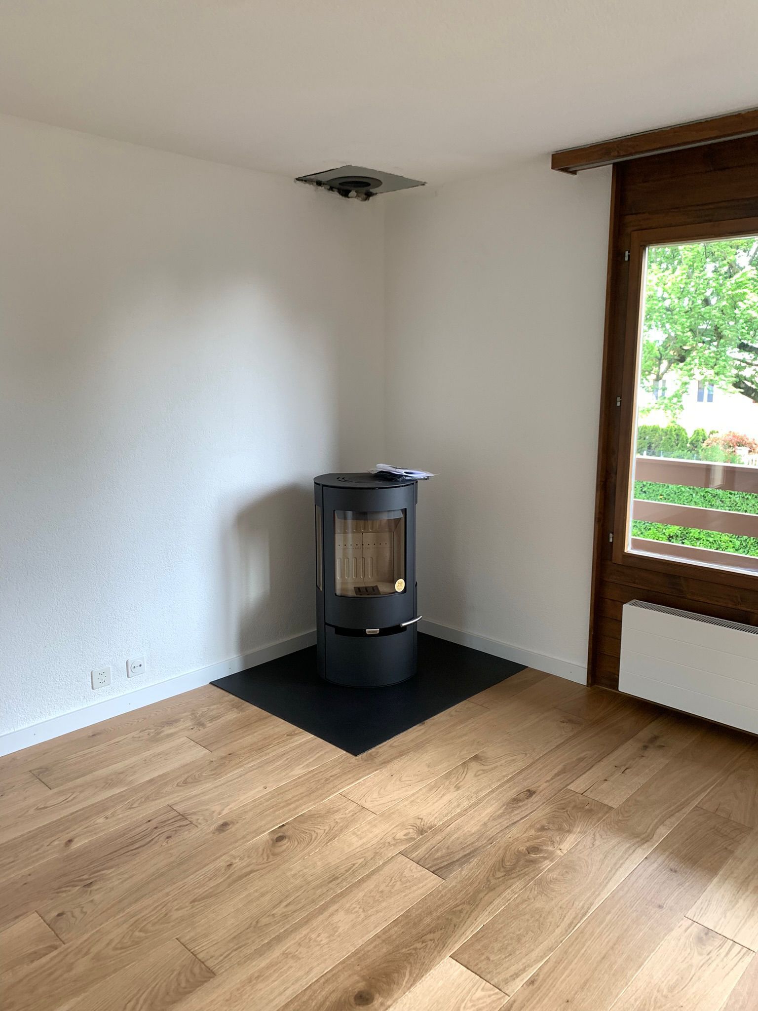 Un poêle à bois cylindrique noir moderne repose sur un socle noir dans un coin d'une pièce au parquet clair et dotée d'une fenêtre.