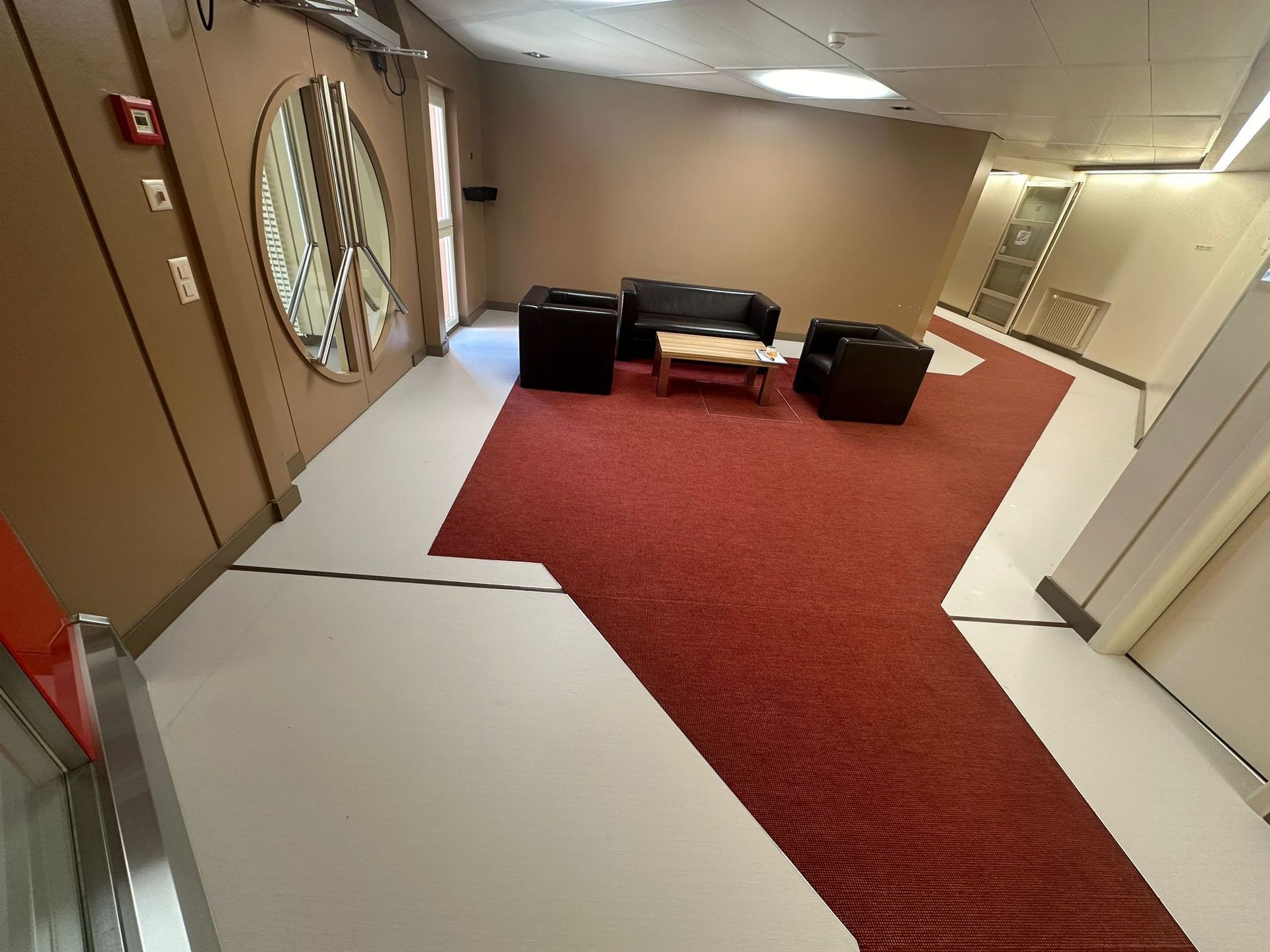 Coin salon avec un canapé foncé et deux fauteuils sur un tapis rouge, sur fond de murs et de sol clairs.