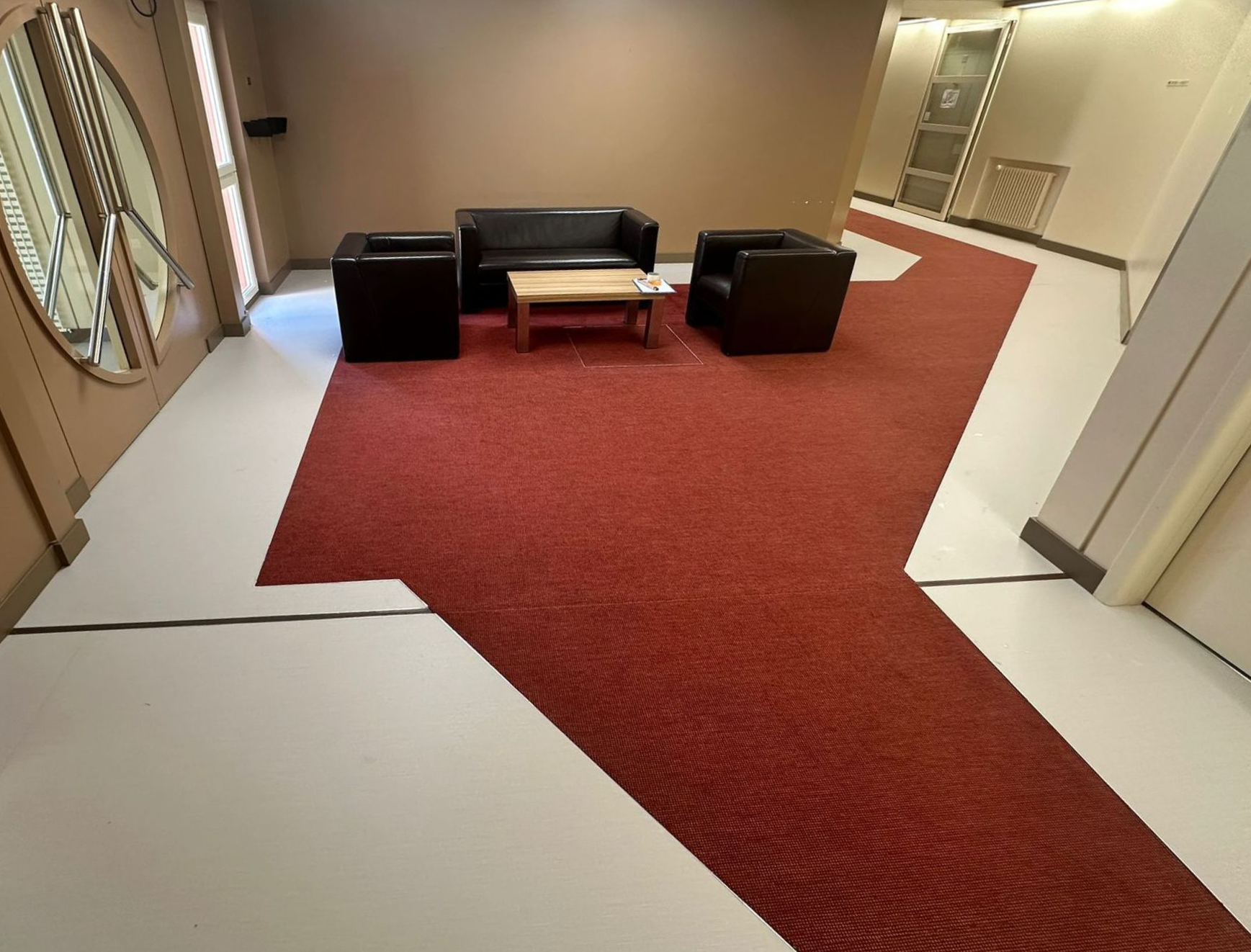 Un hall d'attente avec des meubles noirs sur un îlot recouvert de moquette rouge et entouré d'un sol blanc.
