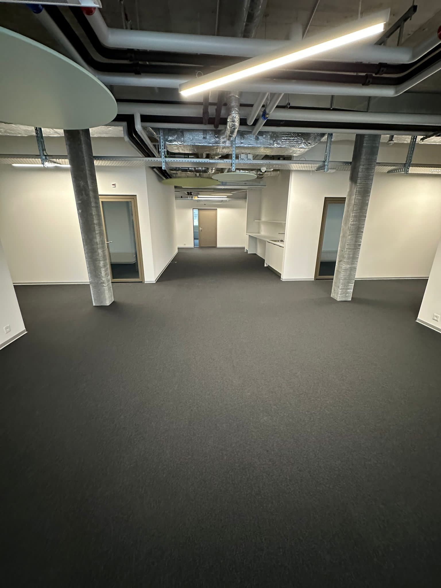 Un couloir de bureau moderne et vide, avec une moquette gris foncé, des tuyaux apparents au plafond et deux piliers structurels proéminents.