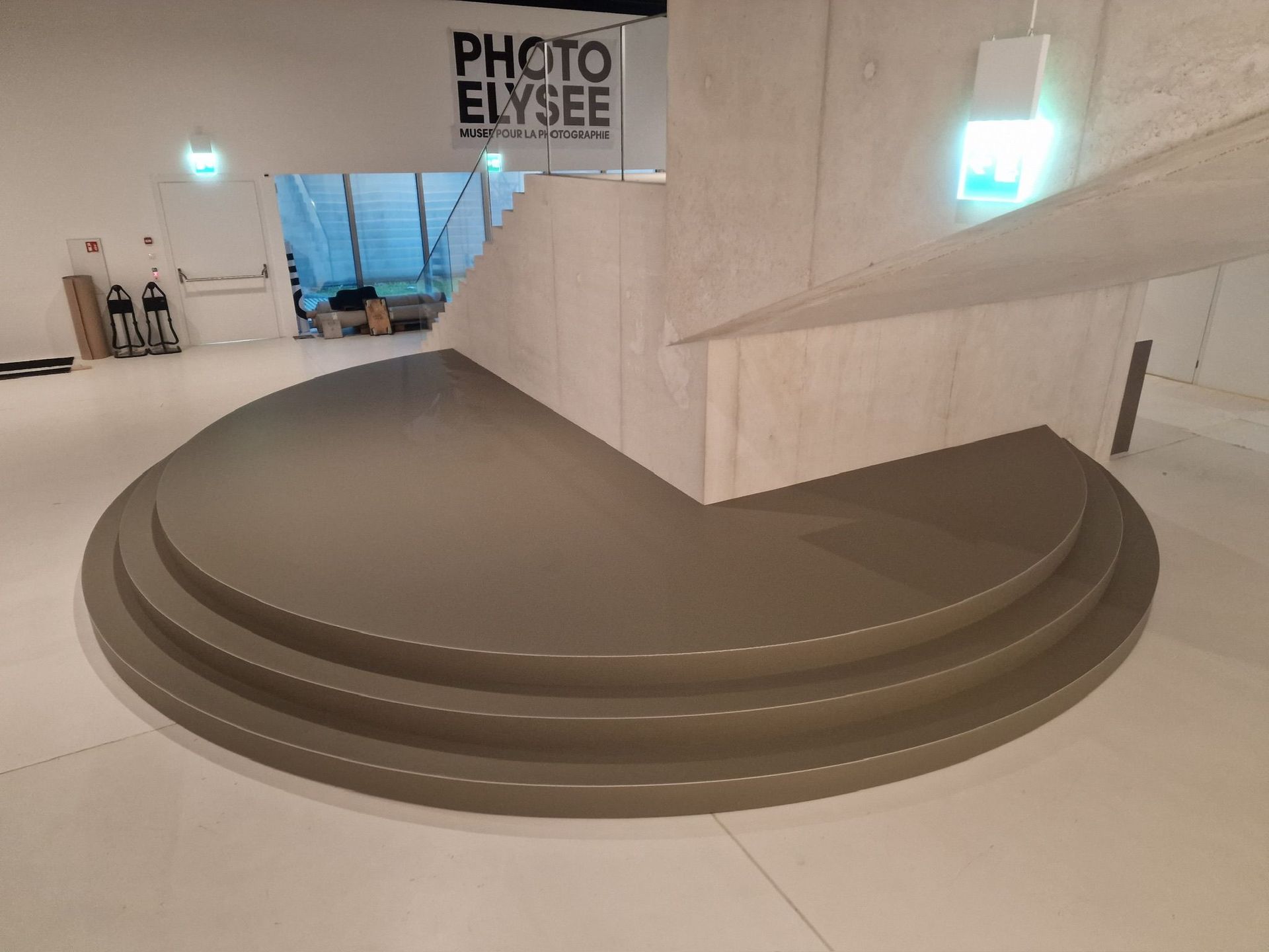 Un escalier semi-circulaire beige, placé au centre d'une galerie intérieure lumineuse et moderne au sol carrelé de blanc.