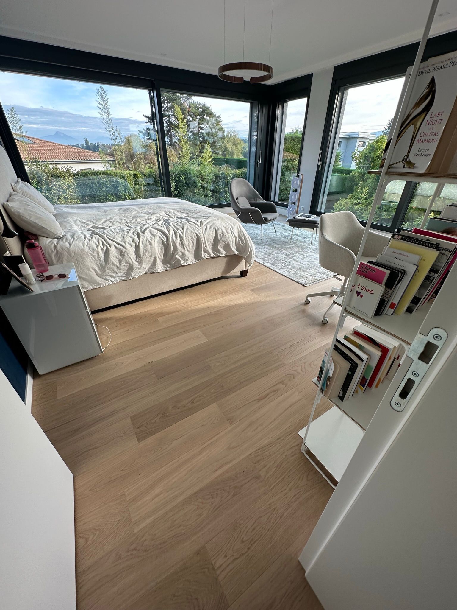 Une chambre lumineuse avec parquet, un grand lit et un bureau face à des baies vitrées donnant sur la verdure.