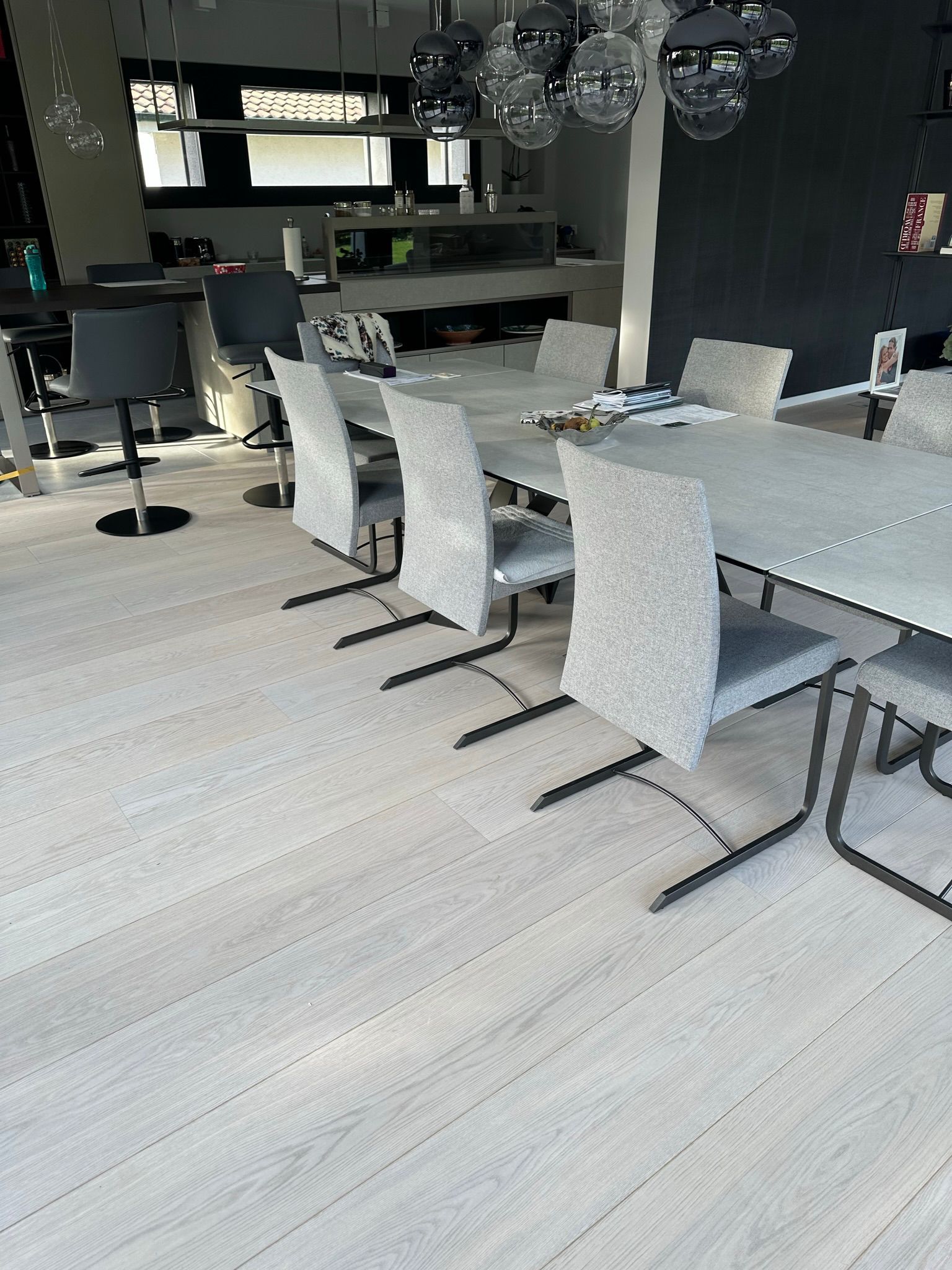 Une salle à manger moderne avec un parquet clair, une longue table et plusieurs chaises grises rembourrées sous des suspensions lumineuses.