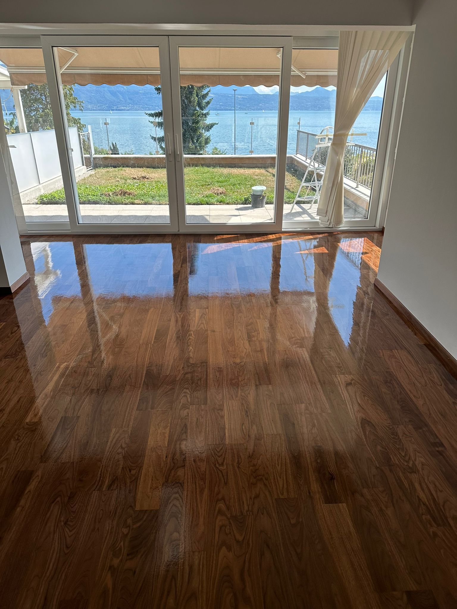 Un parquet brillant reflète une pièce lumineuse et ouverte, avec des portes vitrées donnant sur un jardin et un lac.