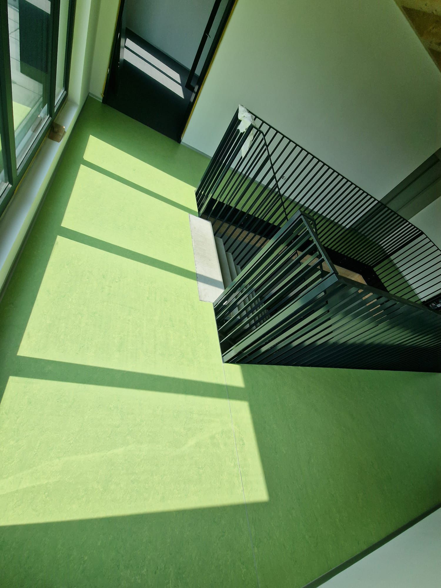 Vue en plongée d'un sol vert clair avec des jeux de lumière du soleil, à côté d'une rampe d'escalier en métal sombre.